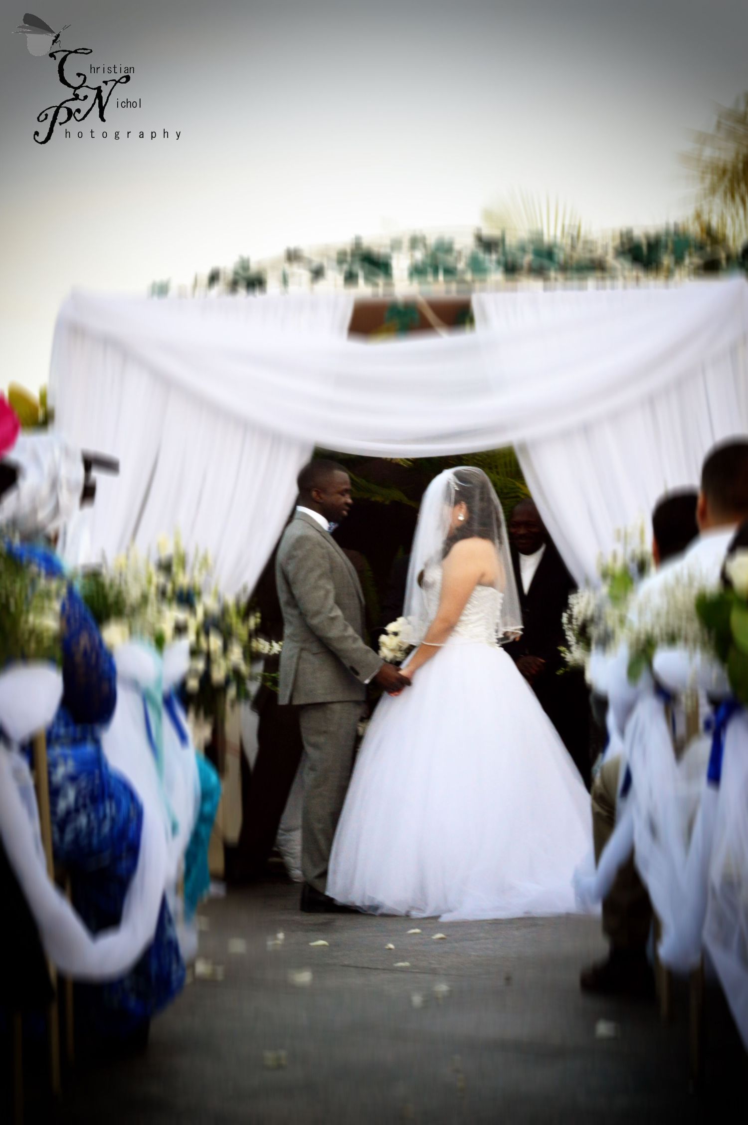 Bride in white gown and groom in gray suit exchange vows under draped white canopy at outdoor wedding ceremony.