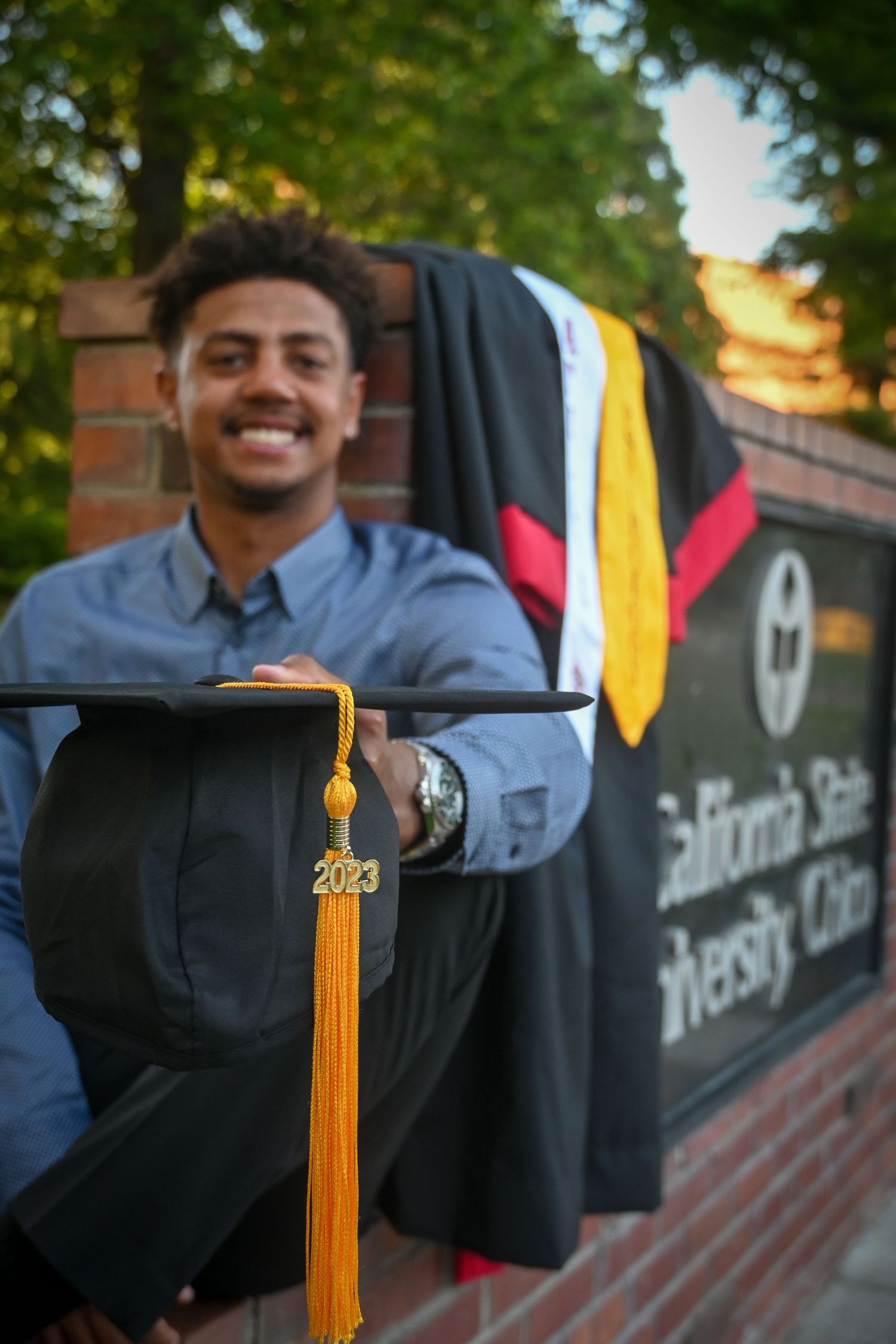 Graduate in blue dress shirt and black graduation gown smiles while holding yellow tassel against brick wall backdrop.