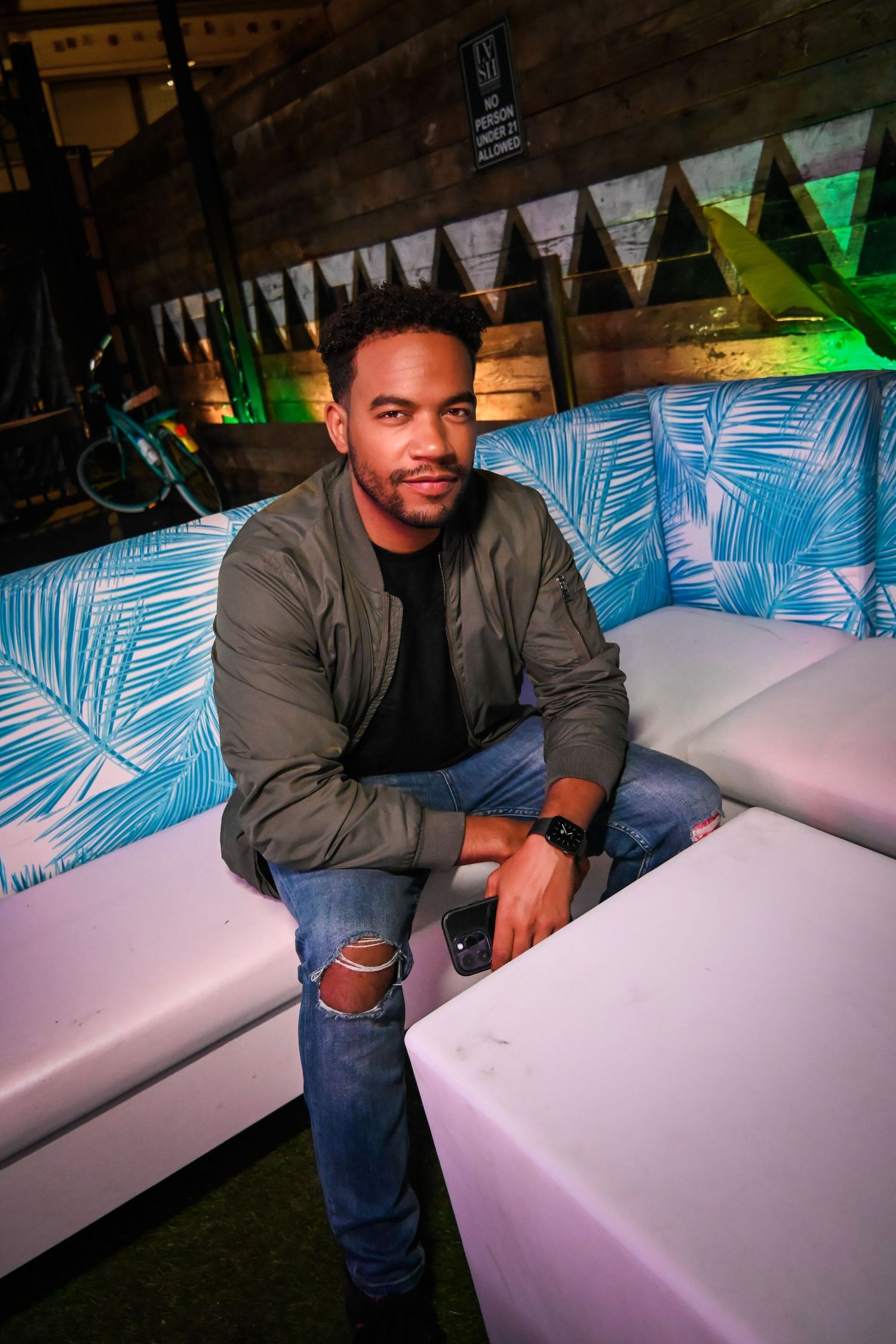 Person in olive bomber jacket and ripped jeans sits on white couch with tropical print pillows at nightclub lounge.