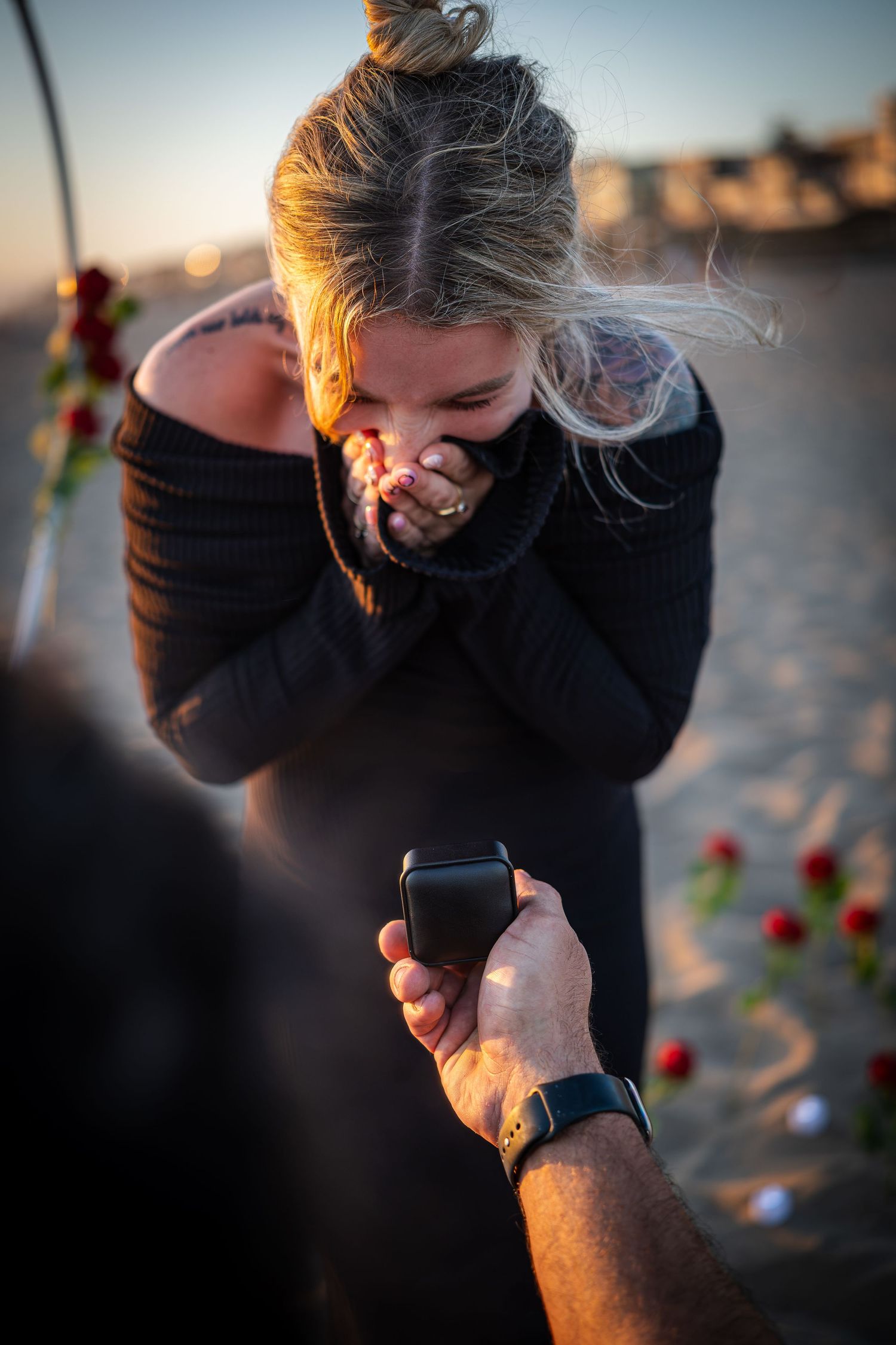 Woman reacts with joy as man proposes with ring box on a beach at sunset with rose petals.