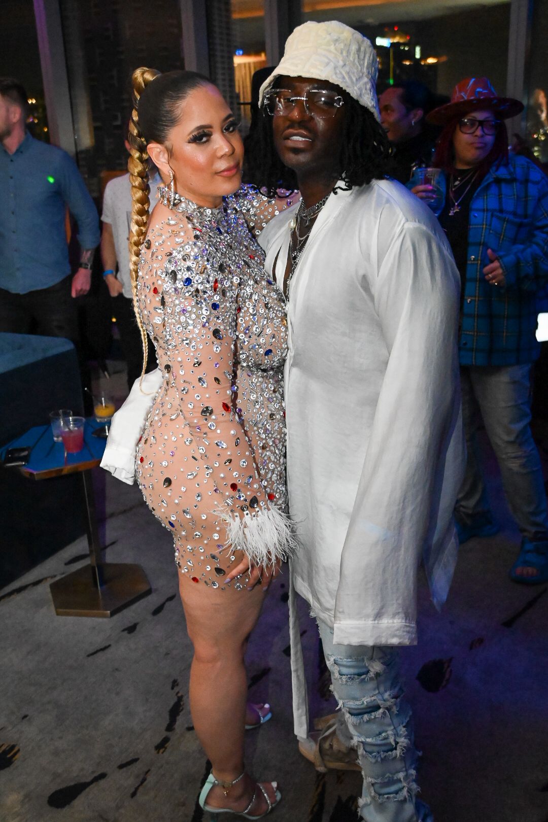 A couple poses together at a nightclub, with one wearing a sparkly sheer outfit and the other in casual white attire.