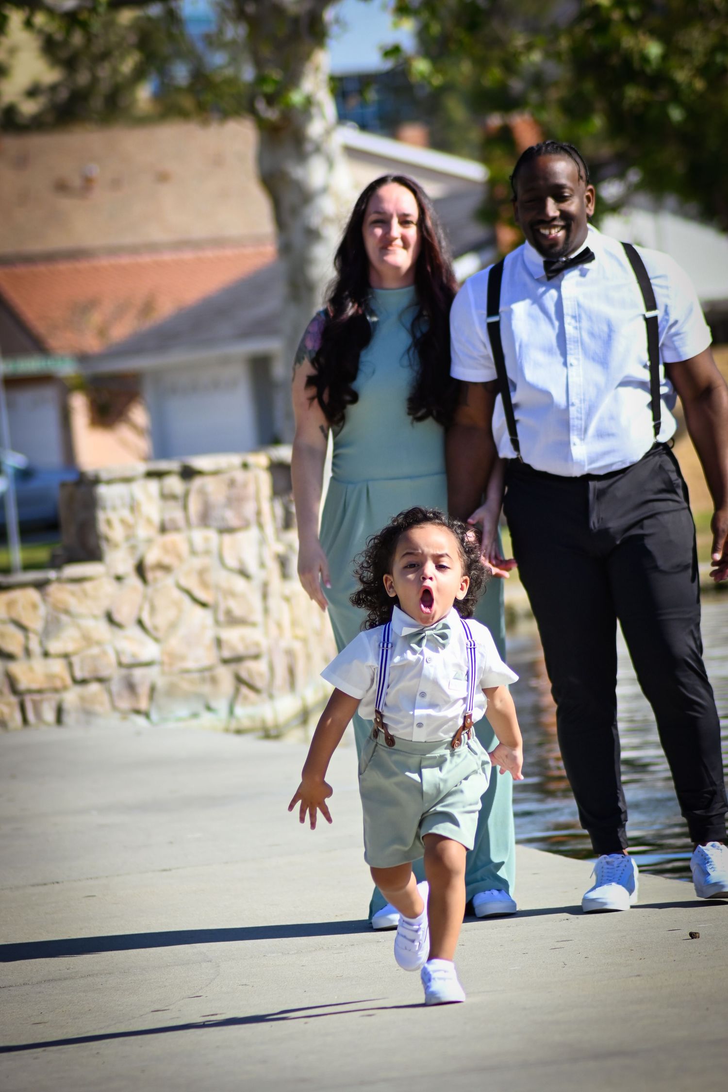 A young child in suspenders and bow tie makes an excited expression while walking with family on a sunny day.