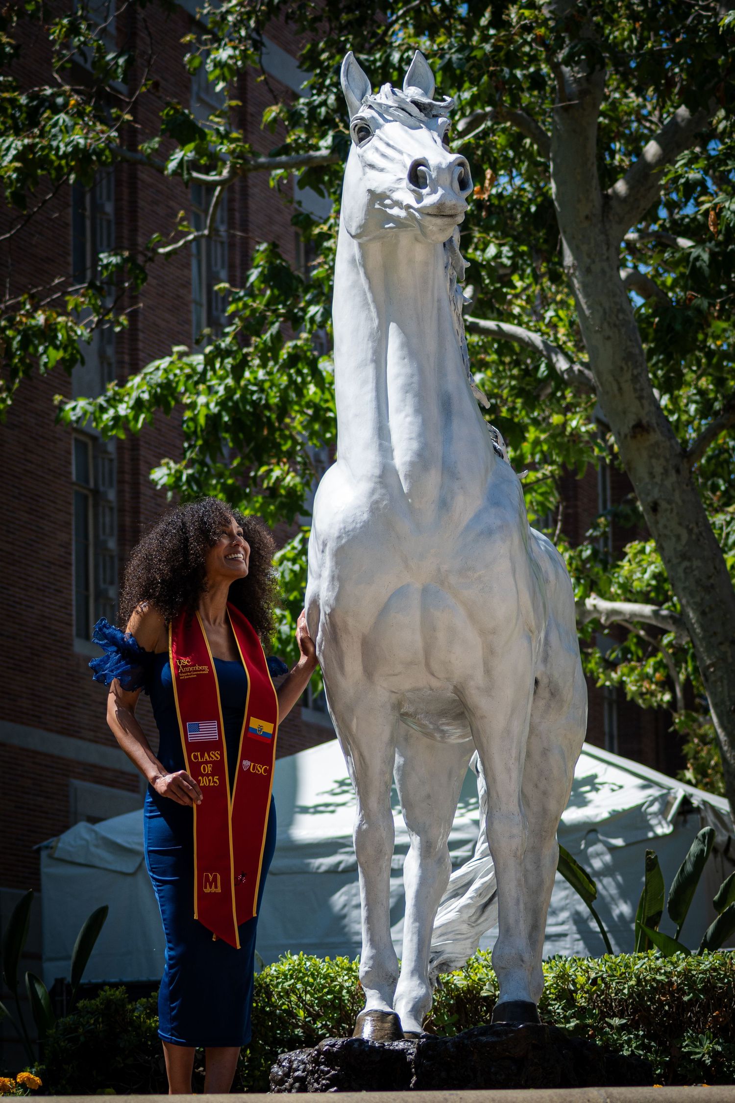 Graduate in blue dress and red stole standing next to large white horse statue outdoors.