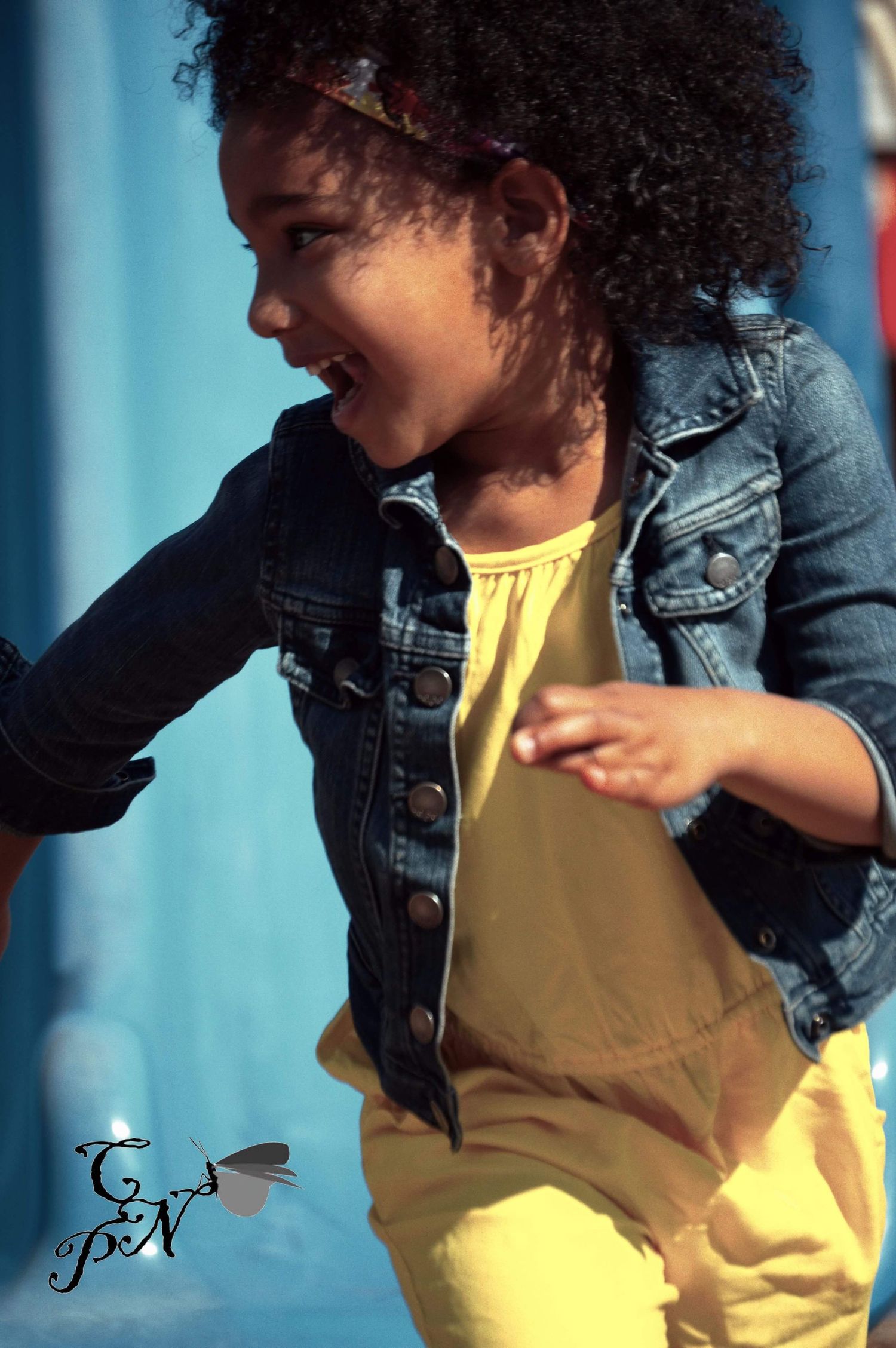 A joyful young girl in a denim jacket and yellow outfit dances outdoors, her curly hair bouncing with energy.