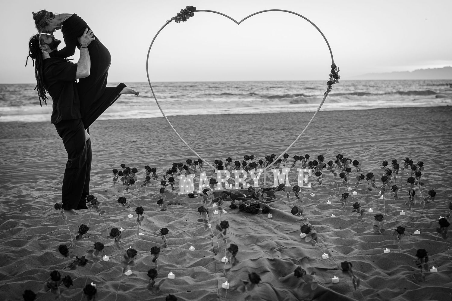 Black and white beach marriage proposal with heart-shaped wire, roses, and MARRY ME sign as couple embraces.