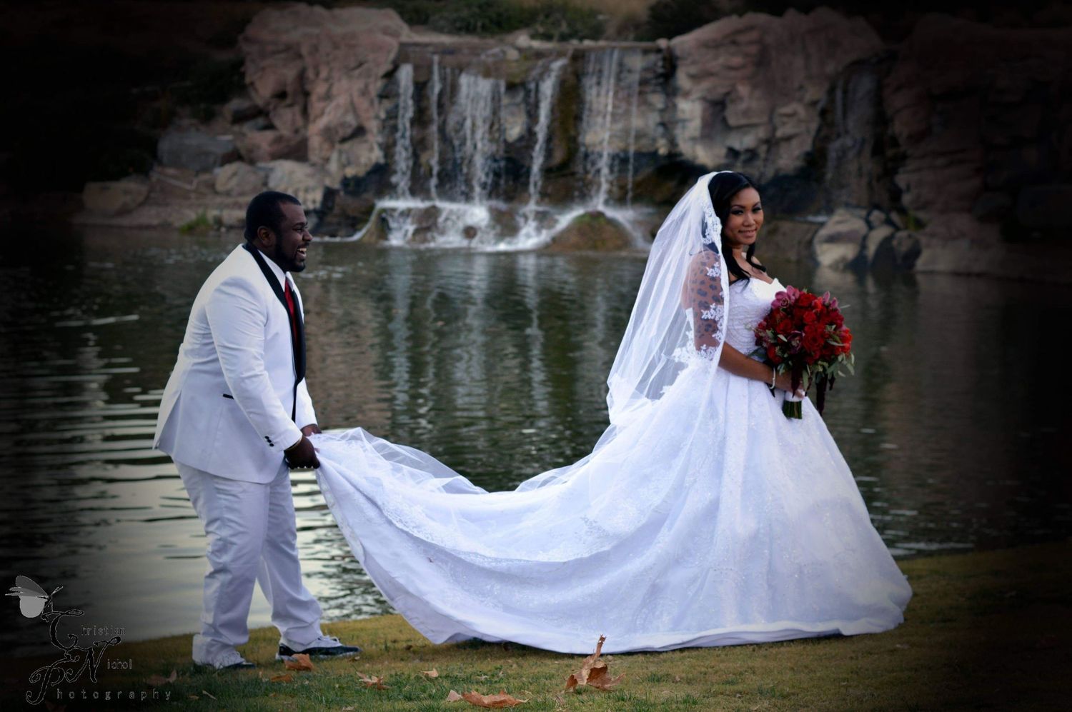 Bride in white gown with long train and groom in white suit pose near a waterfall and pond in an outdoor wedding photo.