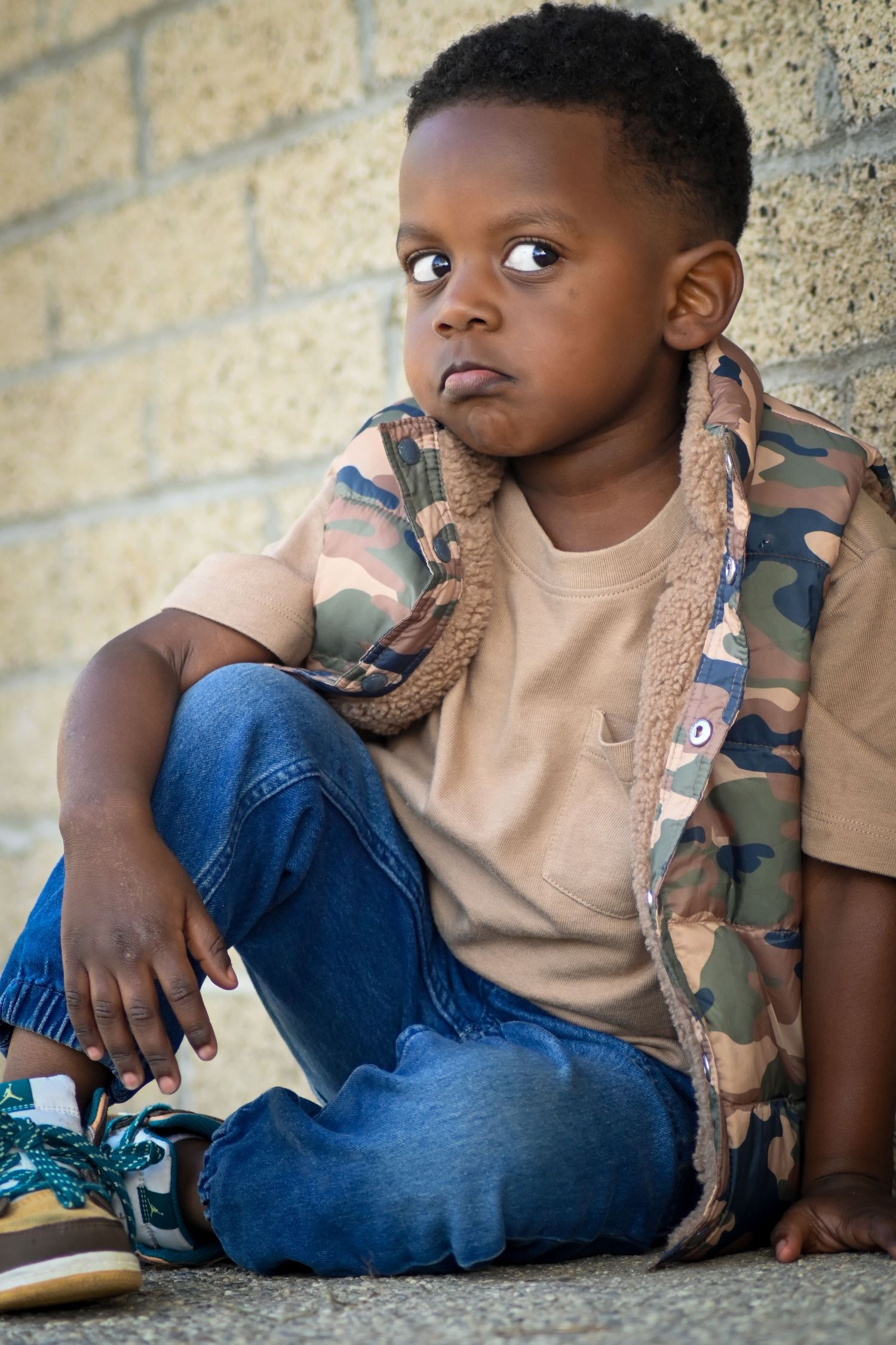 Young Black boy in camo vest and jeans sits against brick wall, looking thoughtful and serious outdoors.