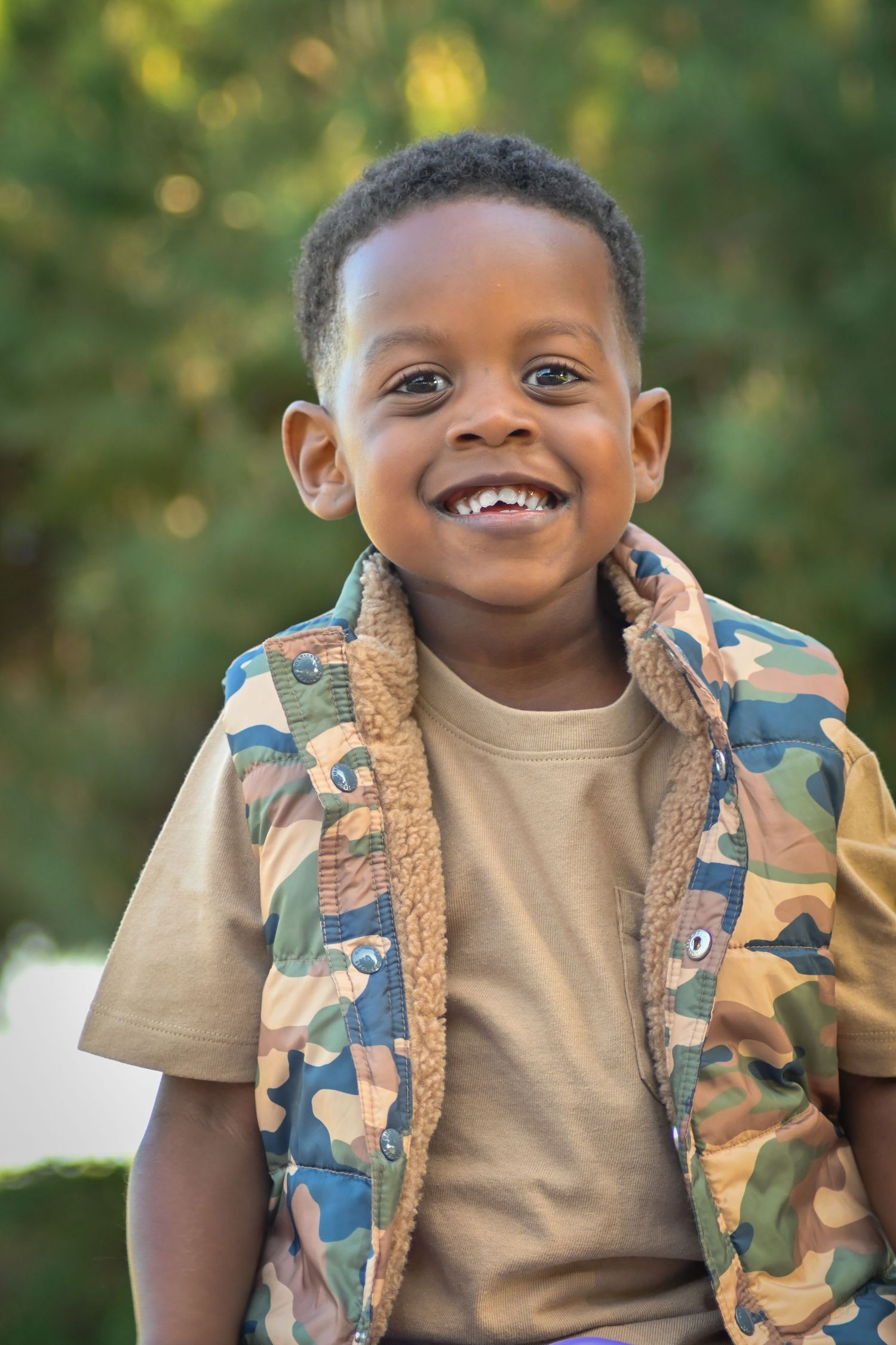 A cheerful young Black boy smiling outdoors, wearing a camo vest over a tan shirt, with green trees in the background.