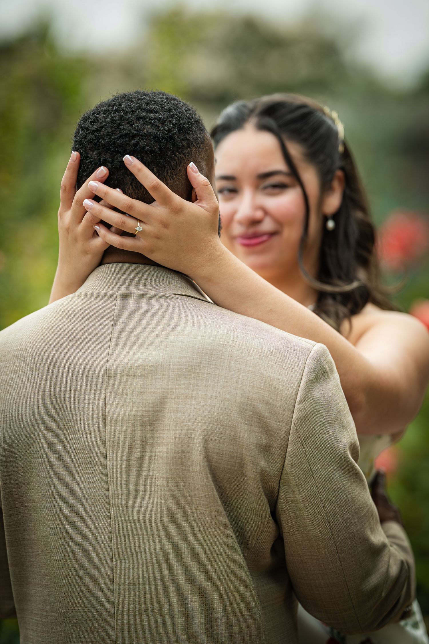 Smiling bride with engagement ring tenderly holds groom's head during outdoor wedding ceremony.