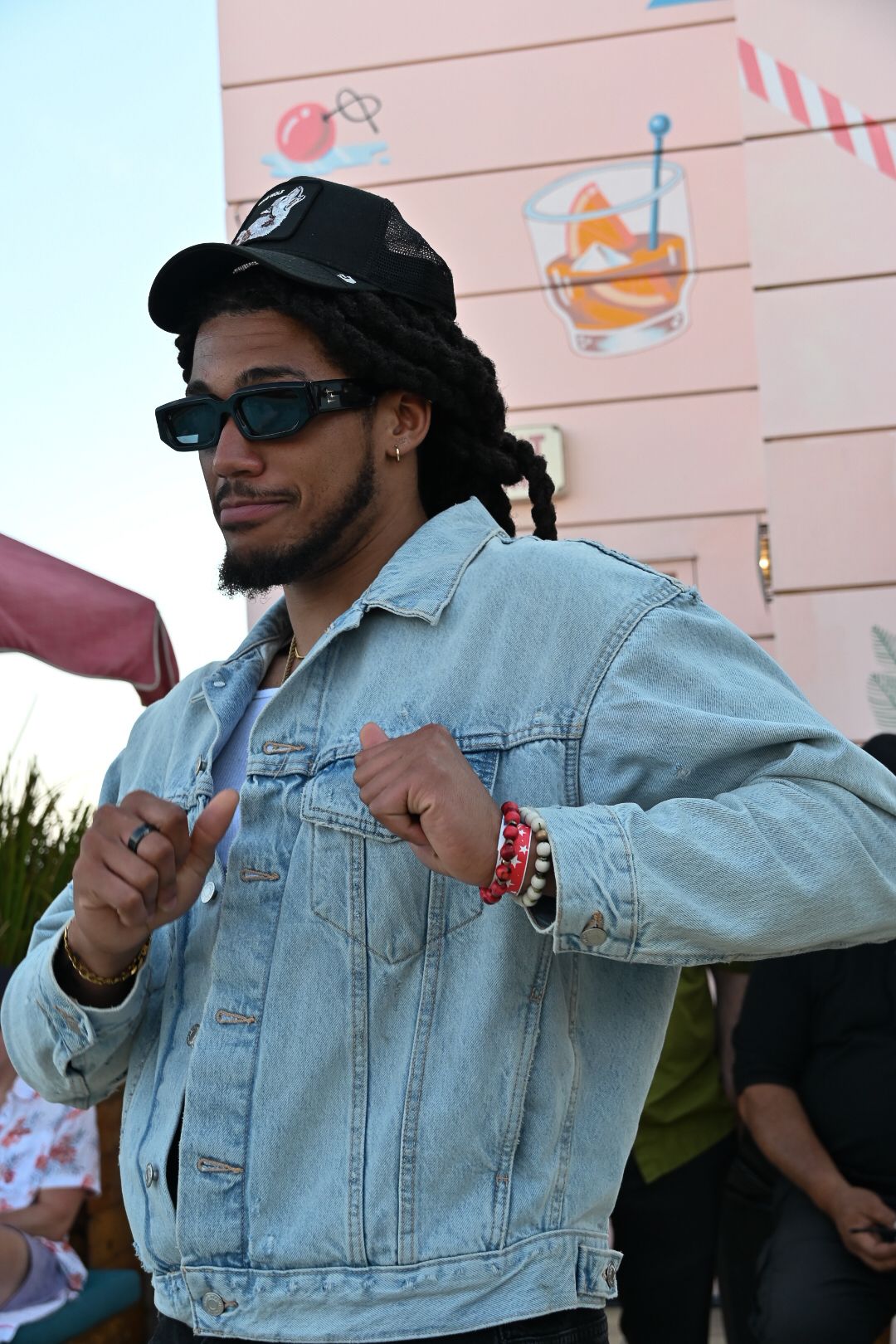 Someone wearing sunglasses and a black cap poses in a light denim jacket against a pink wall with decorative artwork.
