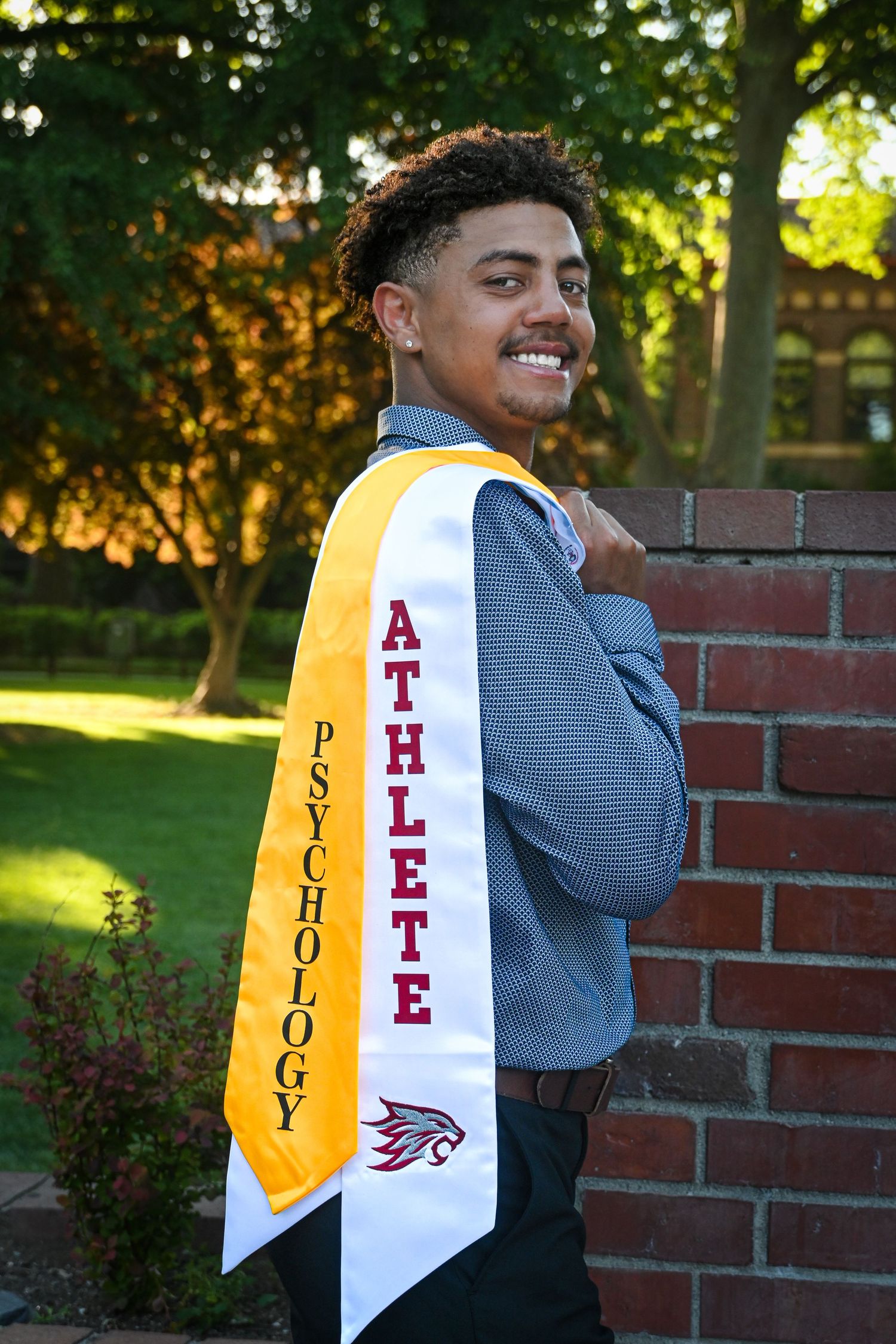 Graduate wearing psychology and athlete graduation stoles poses outdoors by brick wall in natural lighting.