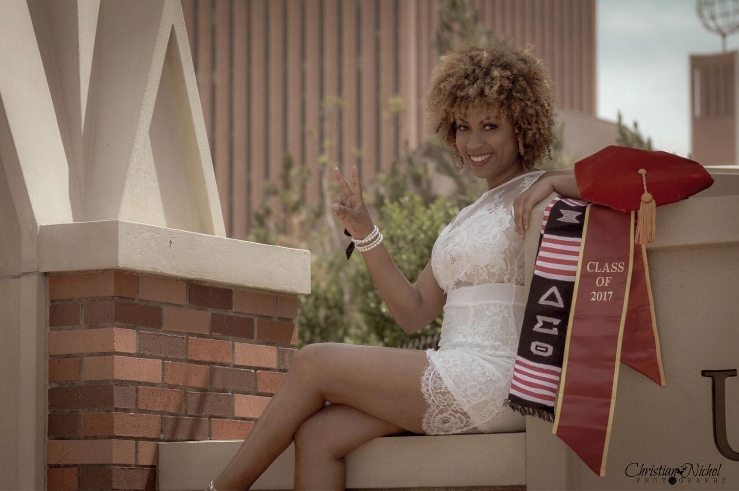 Graduate in white dress sitting on steps with red and gold sash.