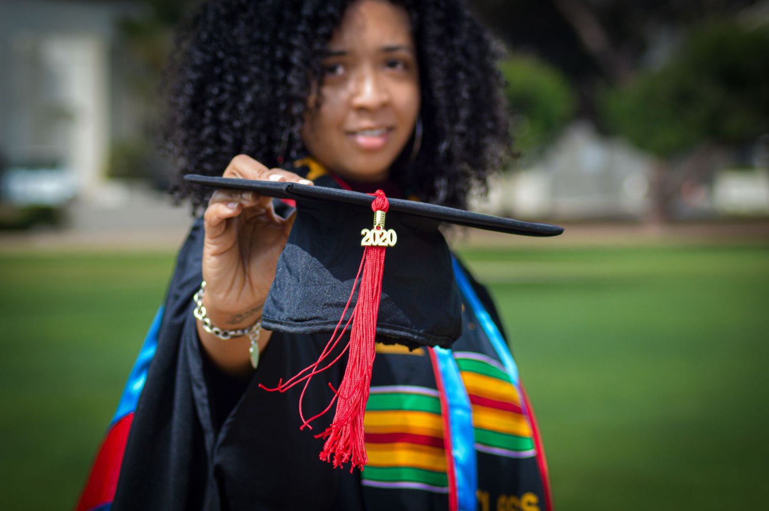 Graduate in cap and gown with rainbow stole stands outdoors during commencement celebration.