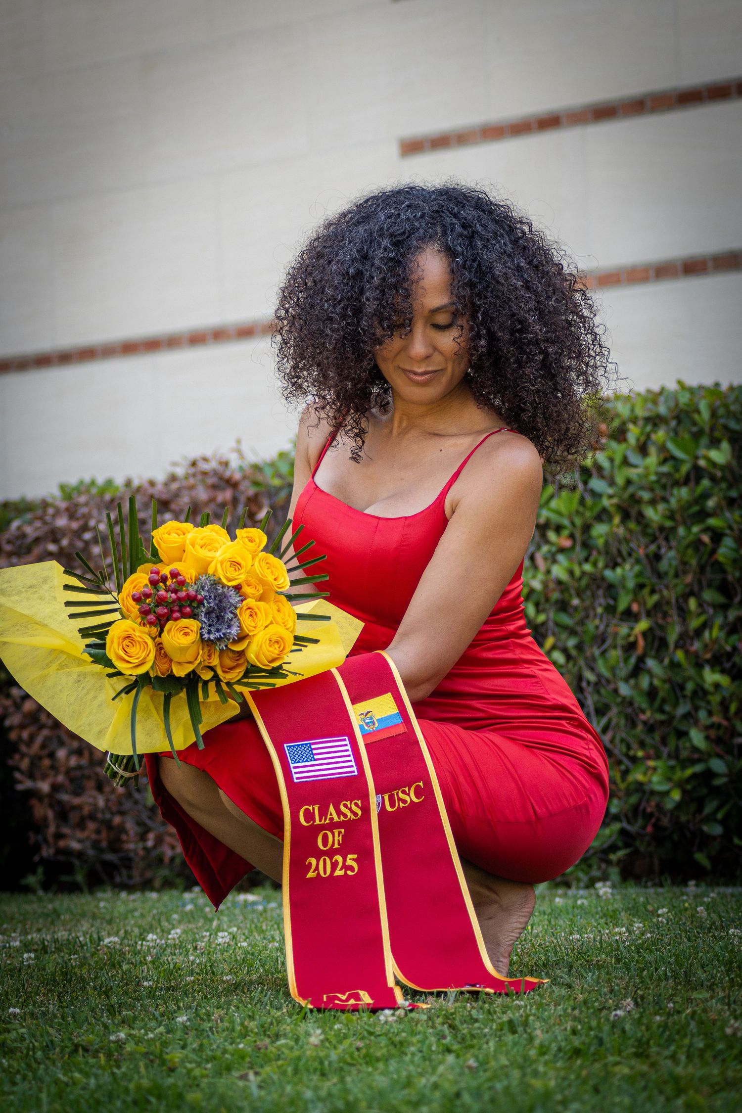 Graduate in red dress poses with sunflowers and graduation stole on grass.