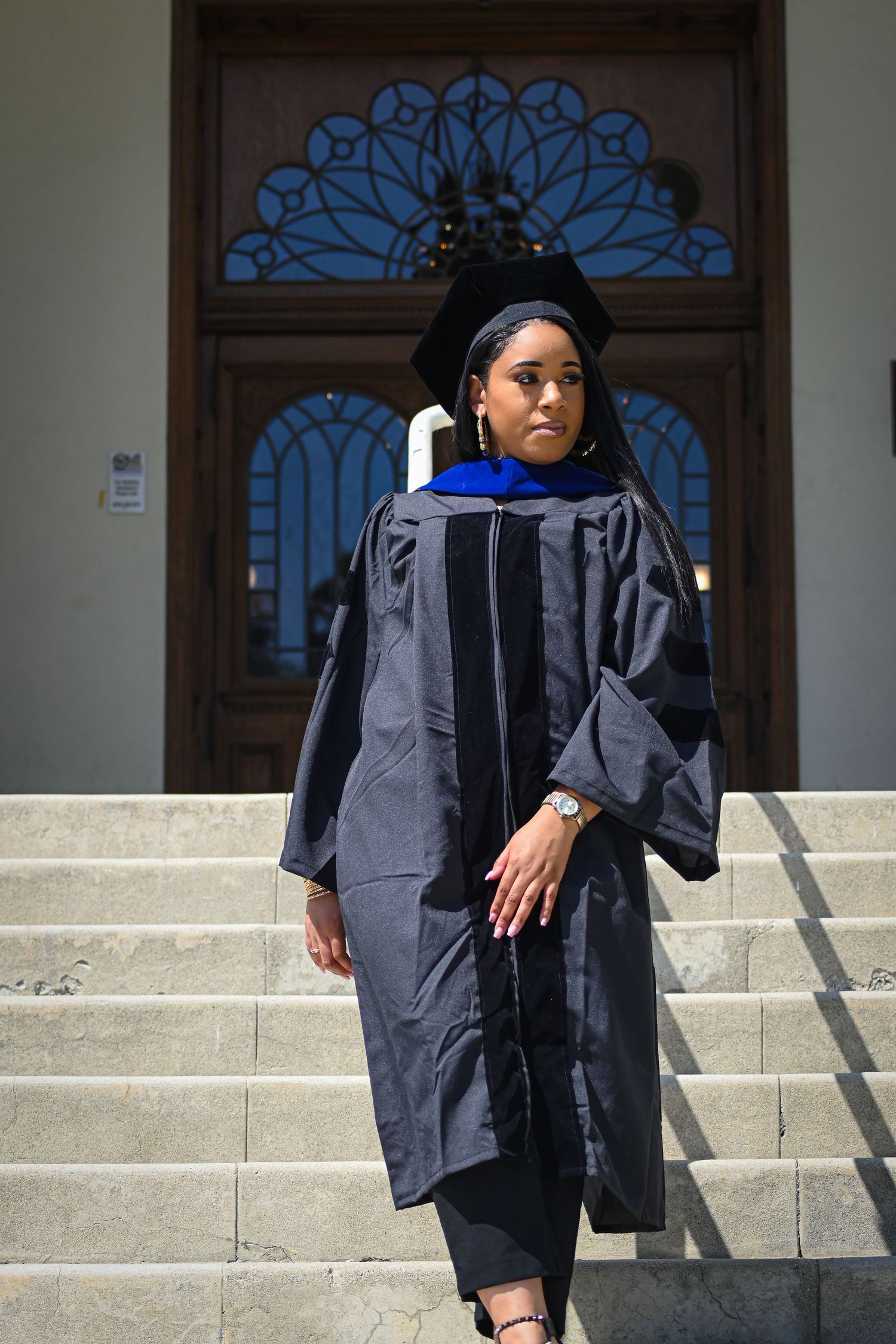 Graduate in academic regalia stands on steps in front of ornate blue window at grand library entrance.
