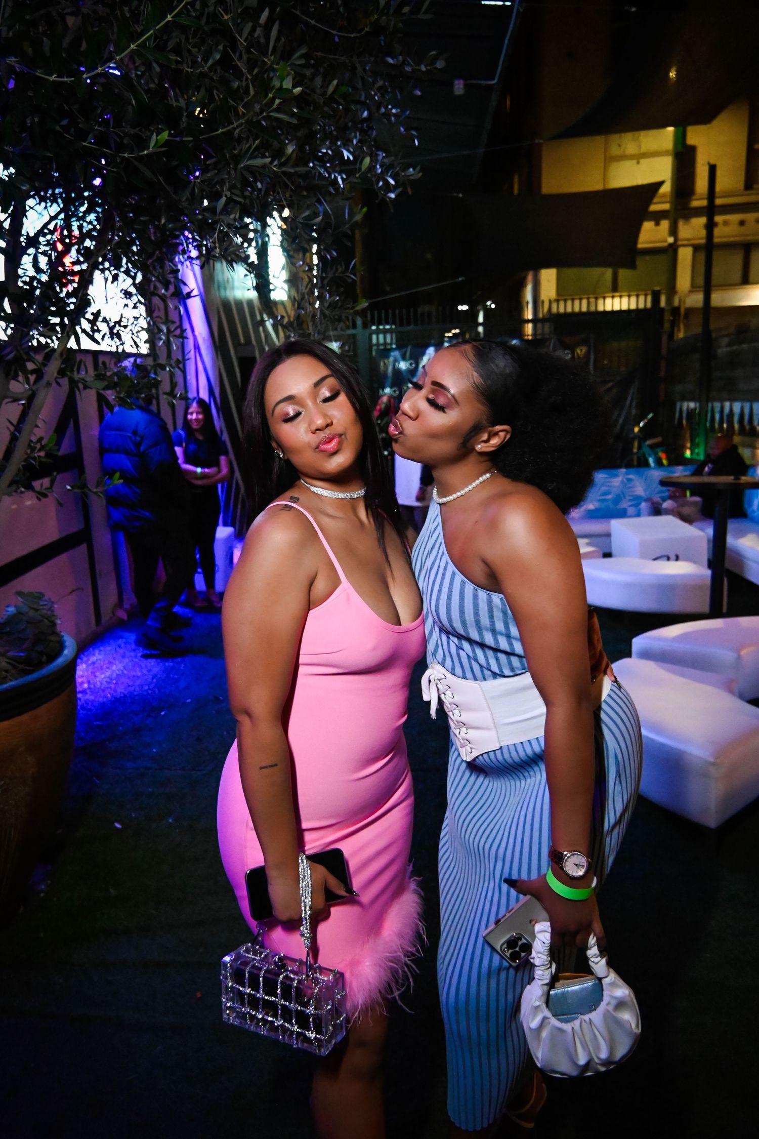 Two people in casual summer outfits enjoying drinks at a nightclub with blue lighting and lounge seating.