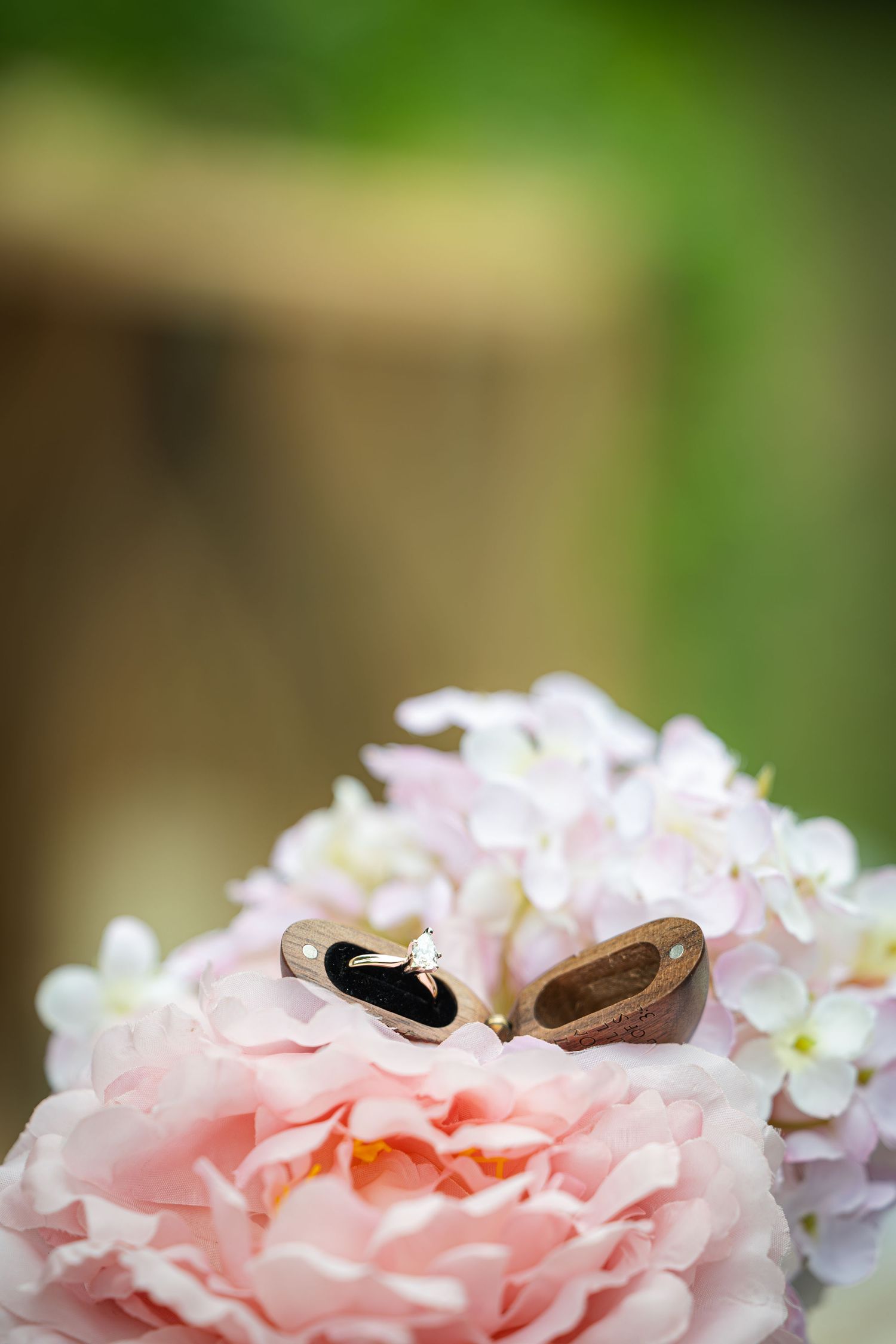 Diamond engagement ring in wooden box on pink peony and white hydrangea bouquet with green bokeh background.