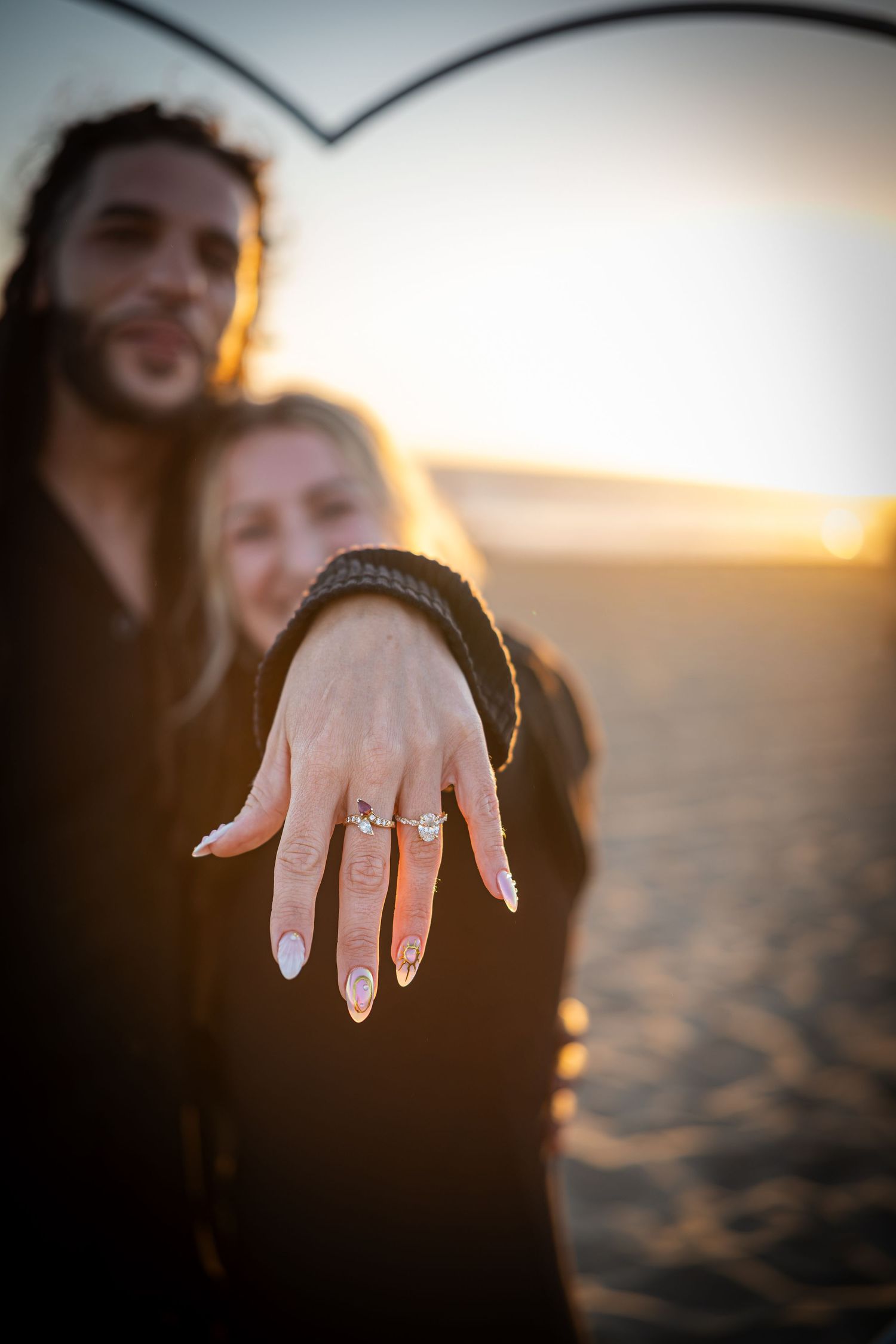 Couple on beach at sunset showing off engagement ring with manicured nails in foreground.