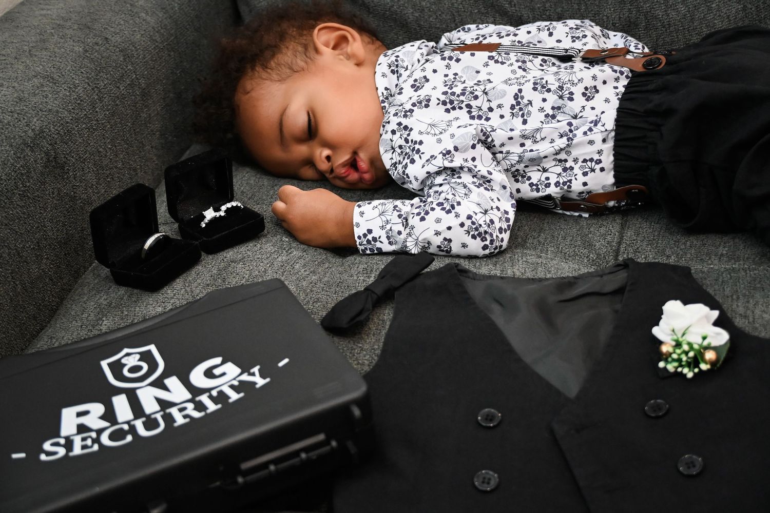 Sleeping toddler boy in tuxedo shirt with suspenders beside Ring Security box, tie, and boutonniere at wedding.