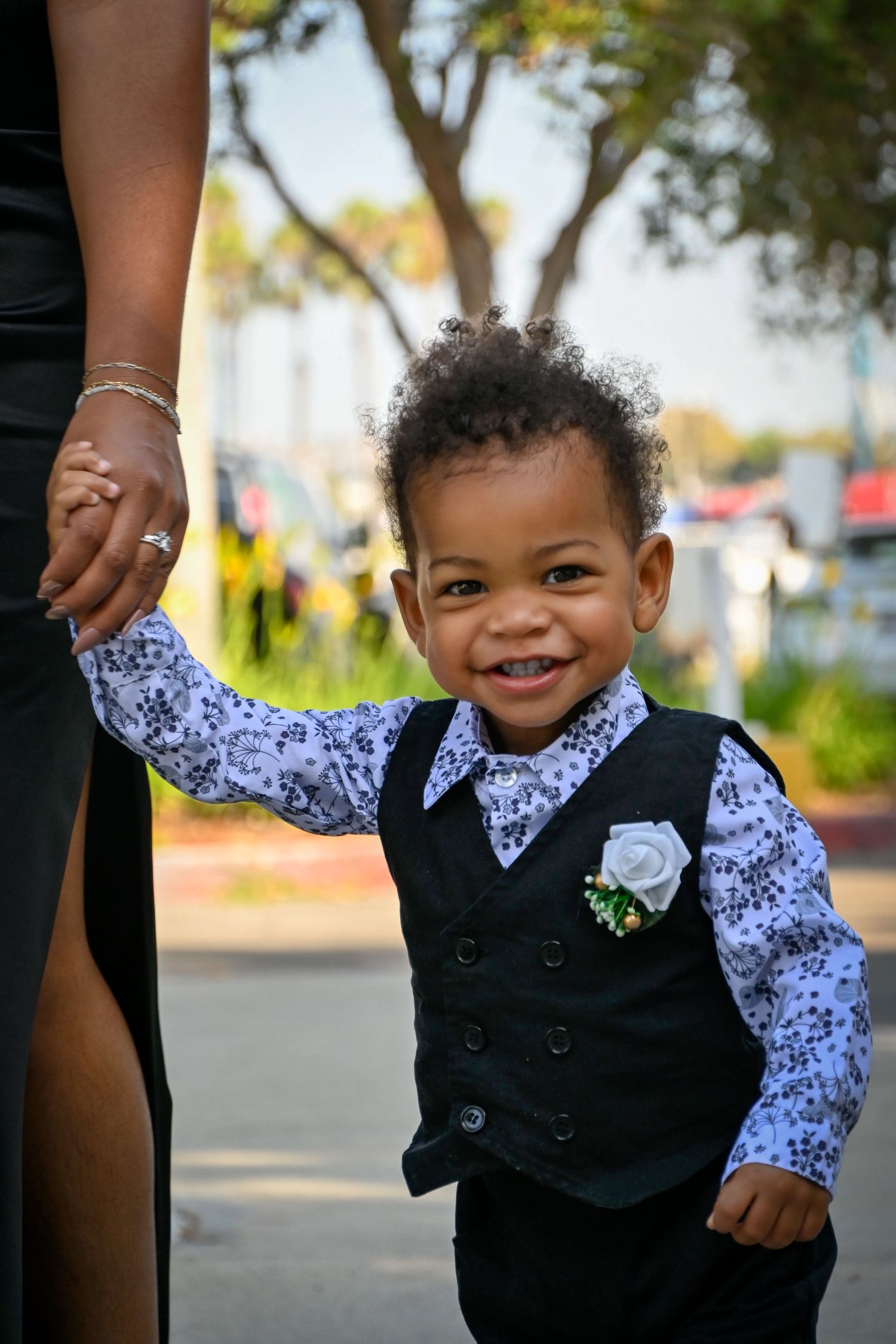 Adorable toddler boy in black vest and floral shirt with white boutonniere smiles while holding an adults hand outdoors.