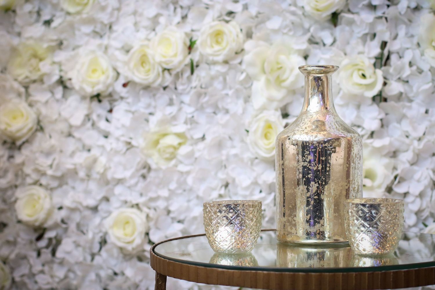 Silver mercury glass vase and votive candle holders on glass table with white rose flower wall backdrop.
