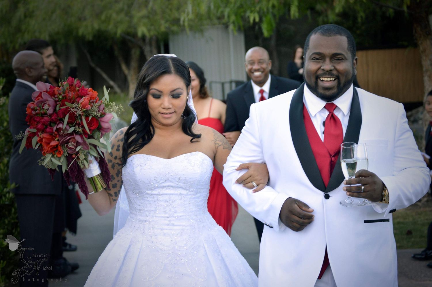 Bride and groom walk joyfully after wedding ceremony, bride holds red bouquet, groom in white tuxedo with red tie.