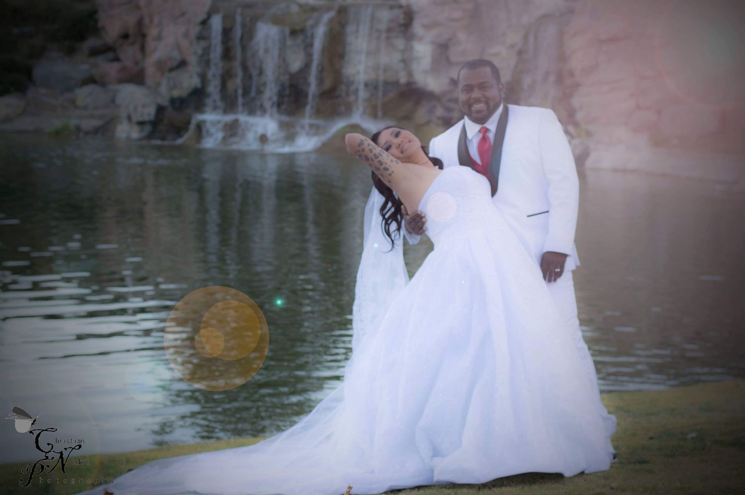 Bride in white gown and groom in white suit pose romantically by a waterfall and pond in an outdoor wedding photo.