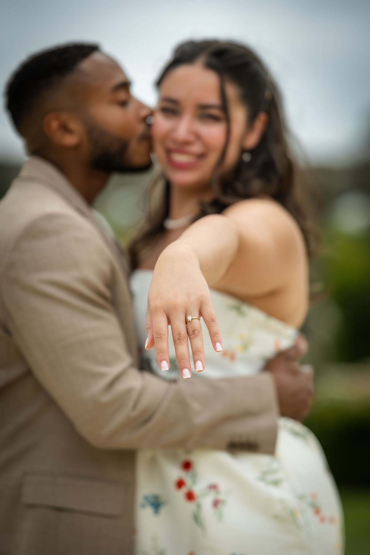 Couple celebrating engagement, woman smiling and showing diamond ring while man kisses her cheek outdoors.