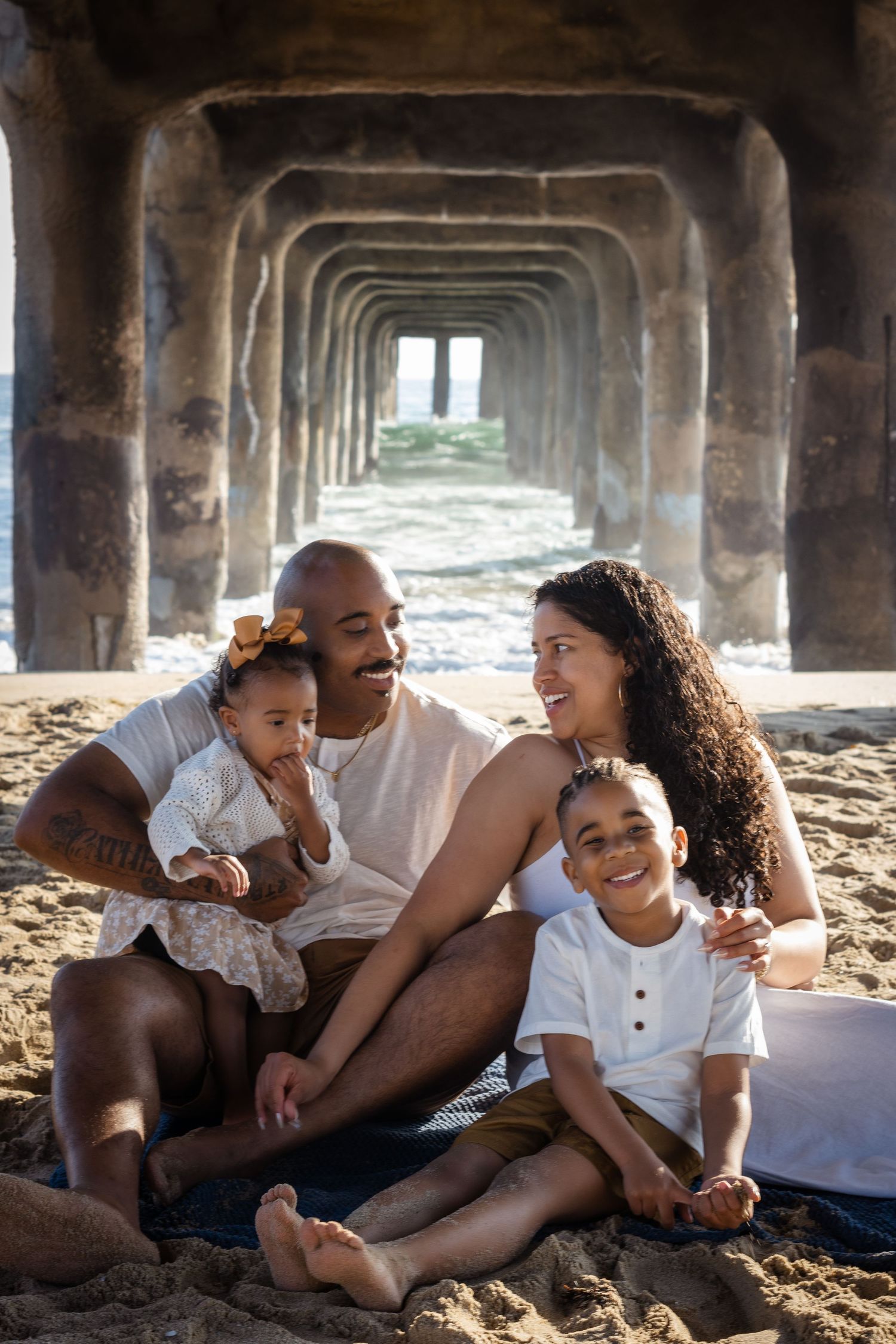 A family relaxes together under a pier at the beach, creating a series of candid moments in black and white.