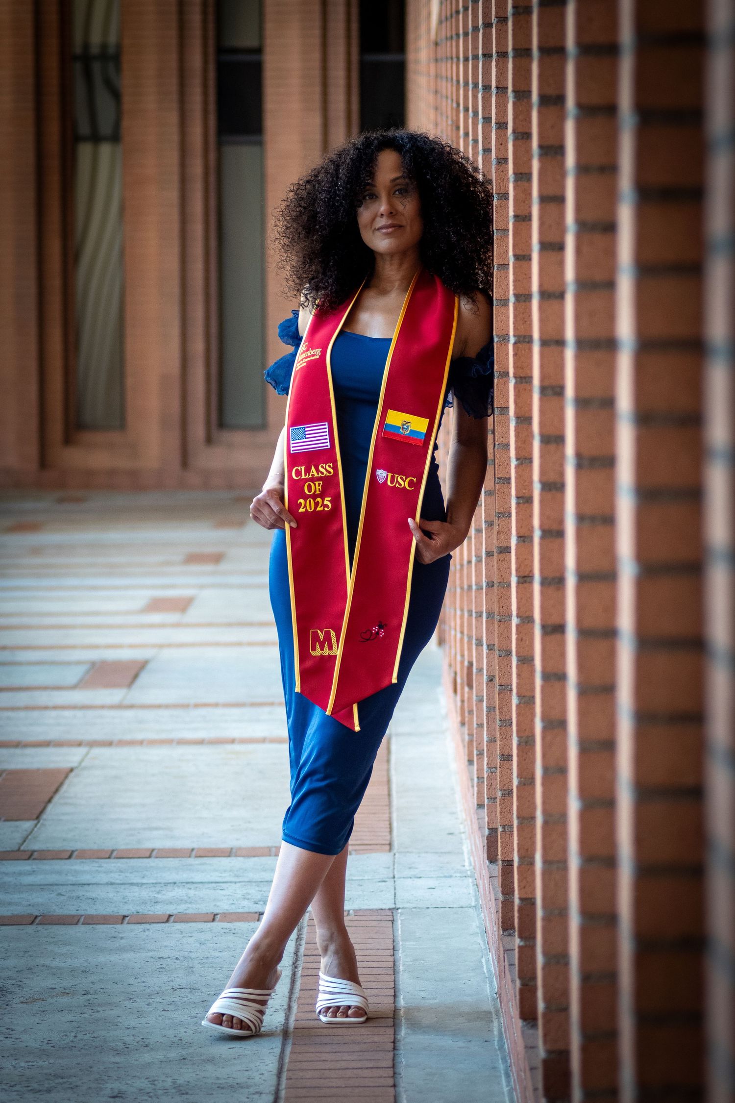 Graduate in blue dress and red sash poses in columned hallway.