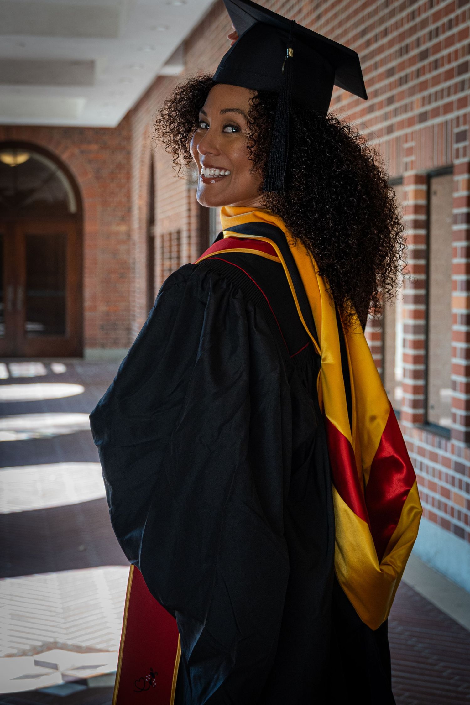 Graduate in cap and gown poses playfully looking over shoulder in brick hallway.