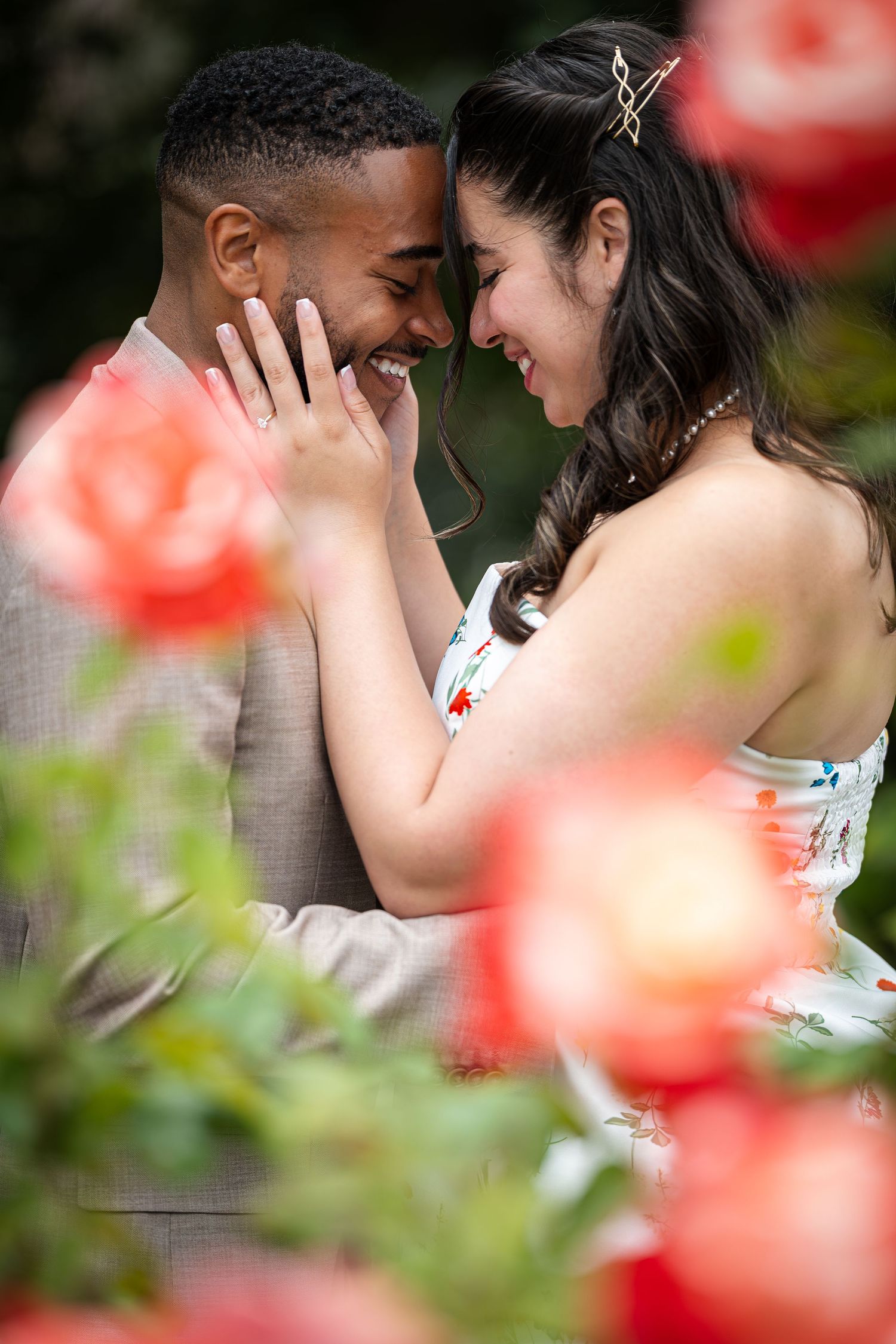 Couple sharing a romantic moment surrounded by blooming red roses, smiling and touching foreheads outdoors.