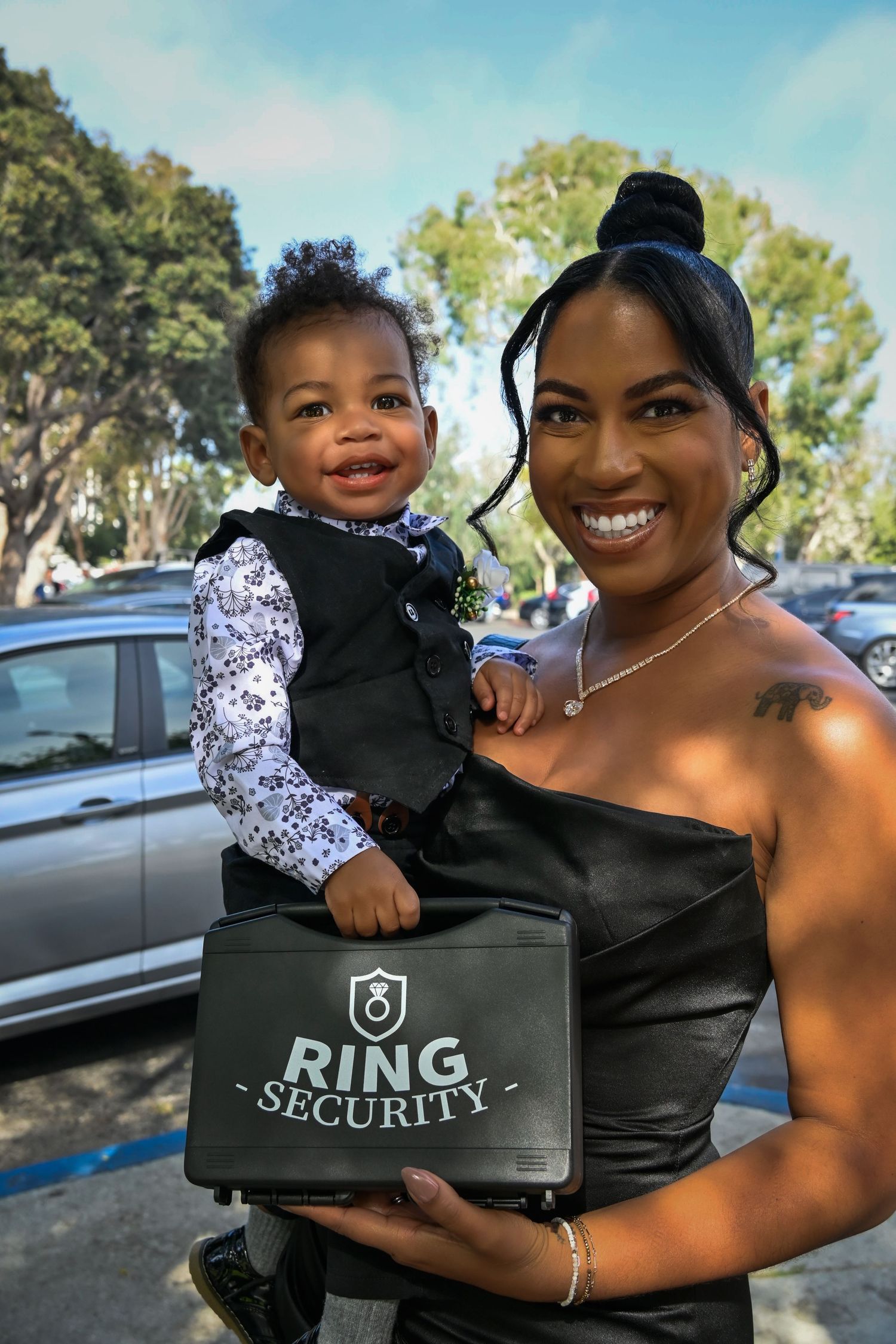 Woman holding baby in vest with Ring Security briefcase at outdoor wedding event.