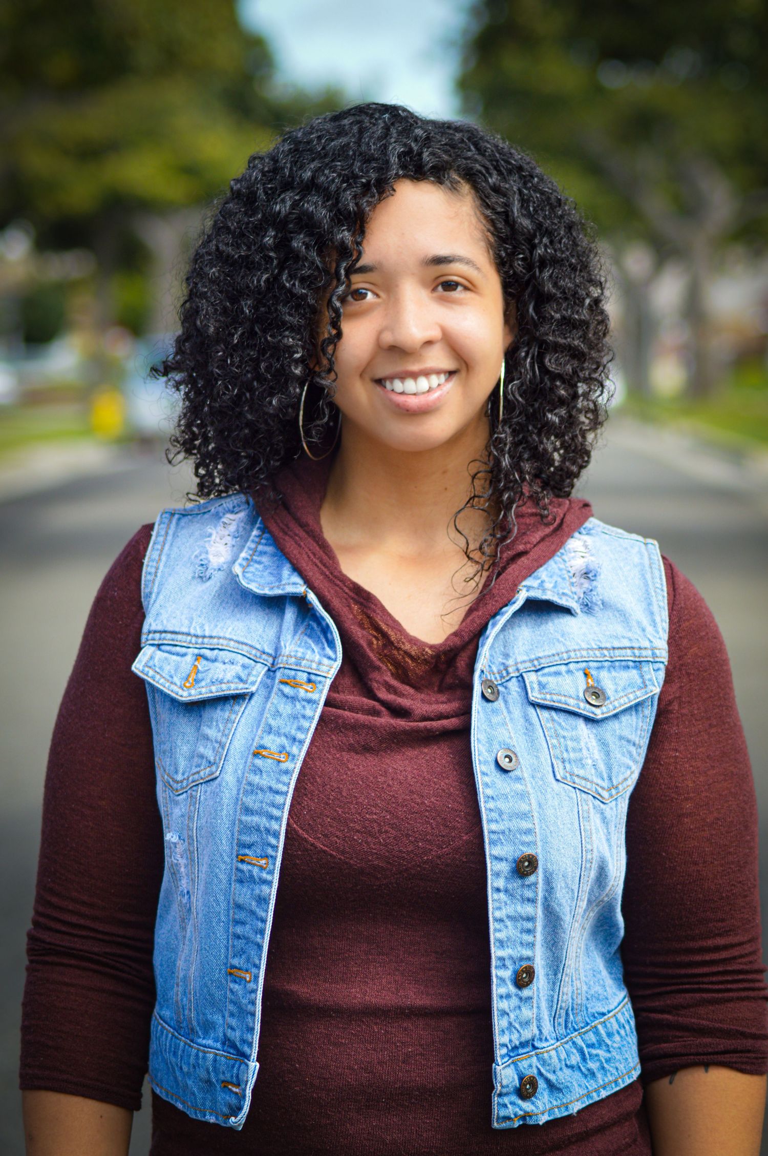 A person wearing a burgundy shirt and denim vest smiles warmly in a series of outdoor portrait photos.