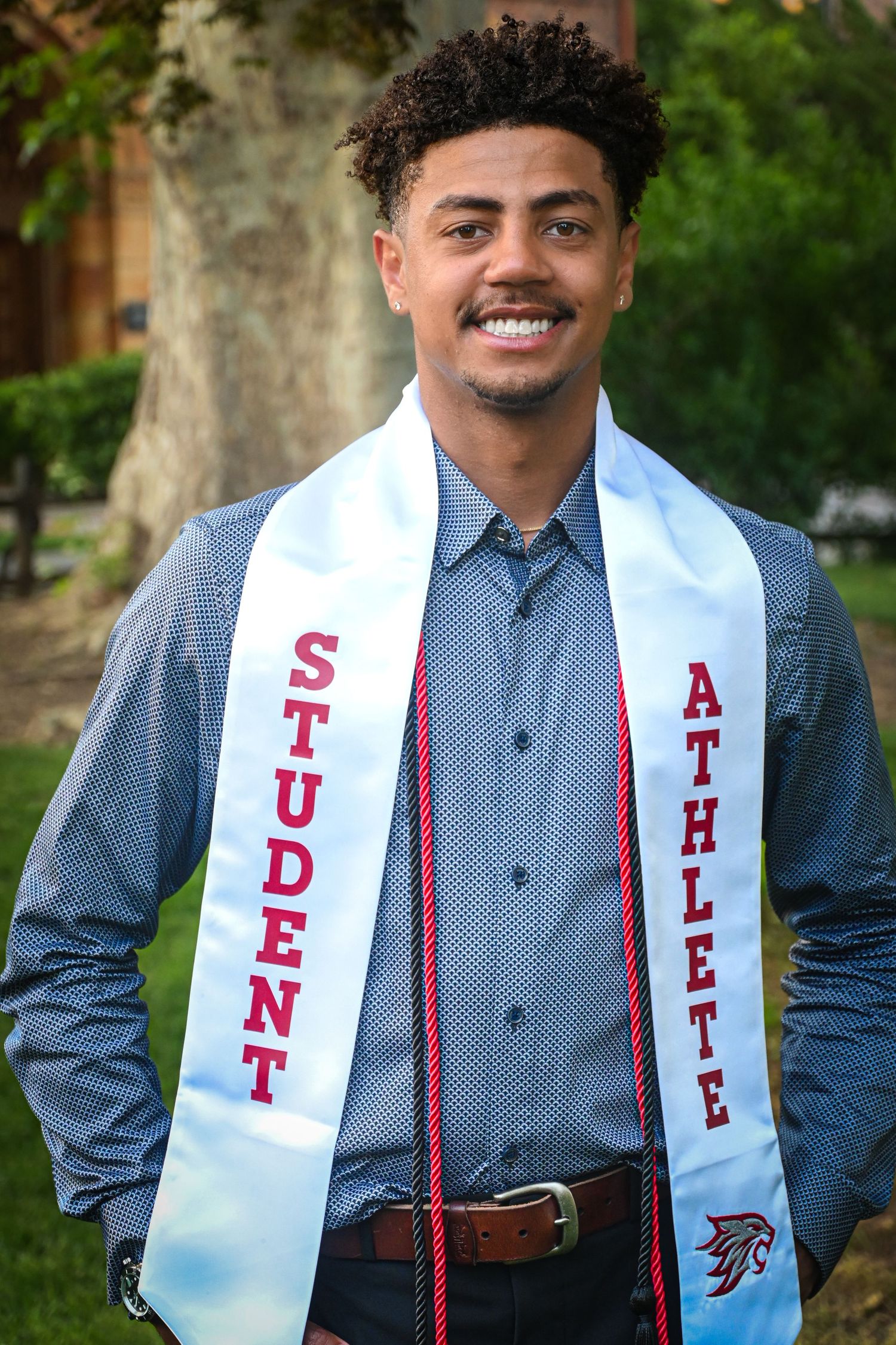 Graduate wearing white sash with Student Athlete text and blue dress shirt smiling outdoors on campus.