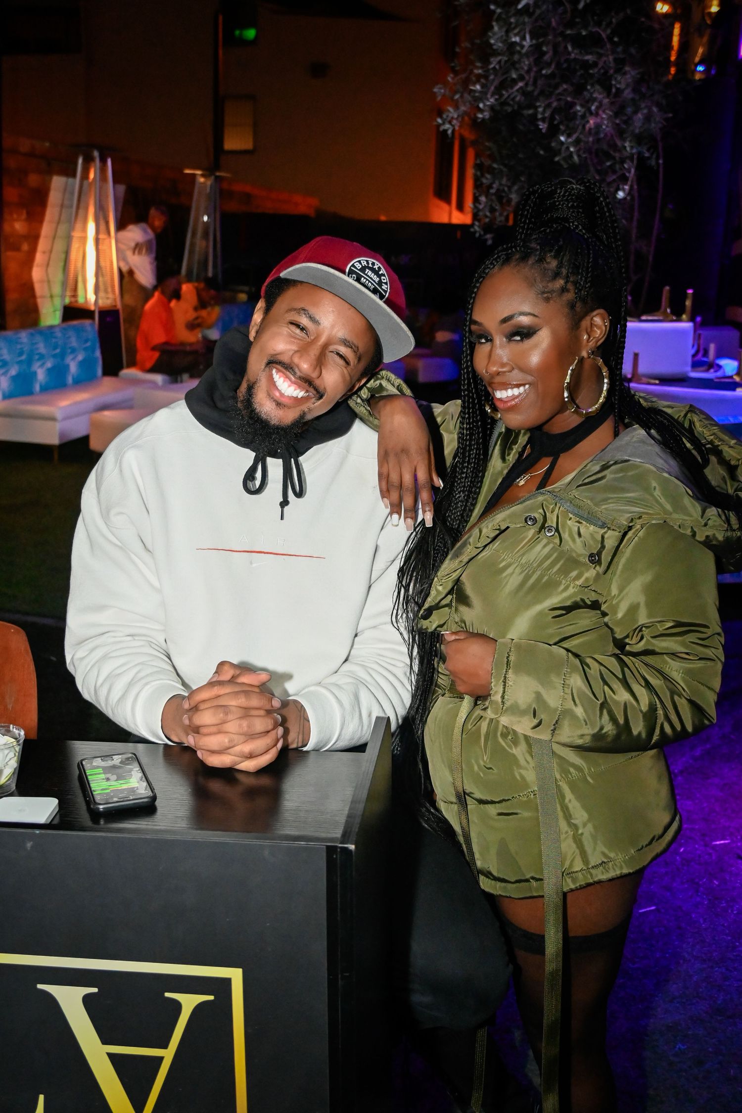Two people pose together at a nightclub table with gold lettering, one wearing a white hoodie and one in an olive jacket.