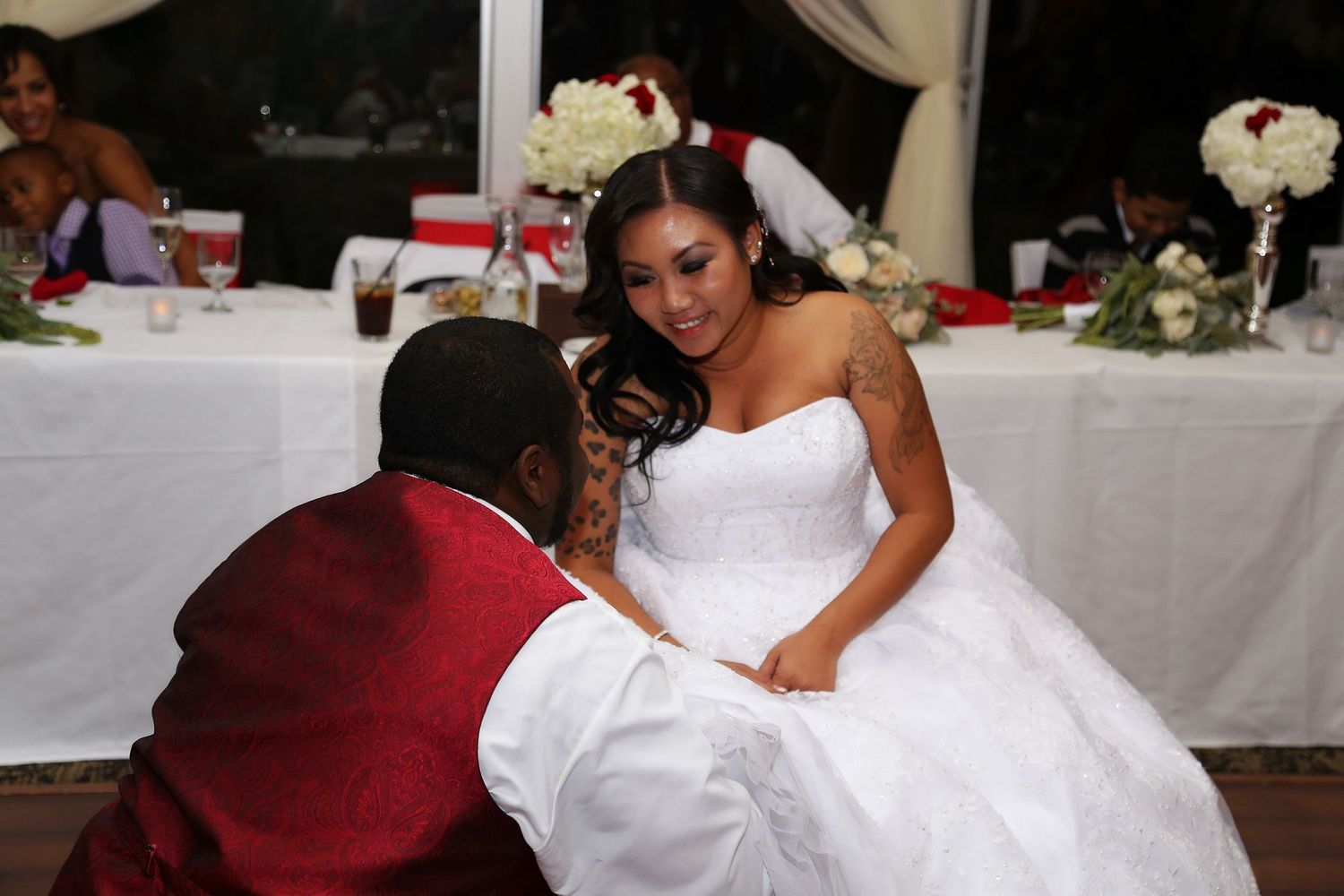 Groom kneeling before bride in white strapless gown at elegant wedding reception with red and white decor.