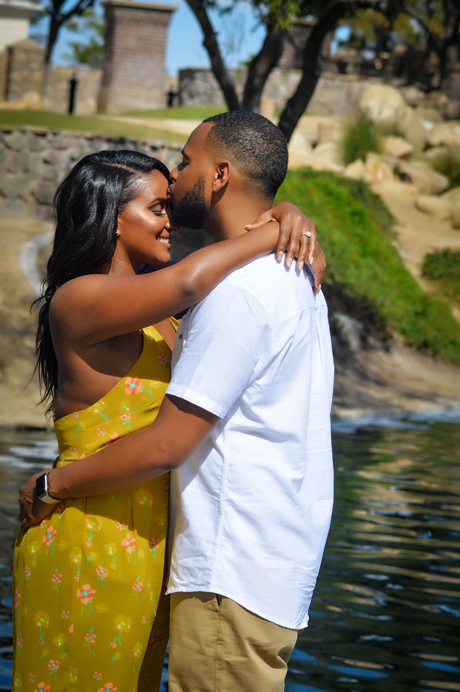 A couple embracing romantically near a waterfront, woman in yellow floral dress smiling as man kisses her cheek.