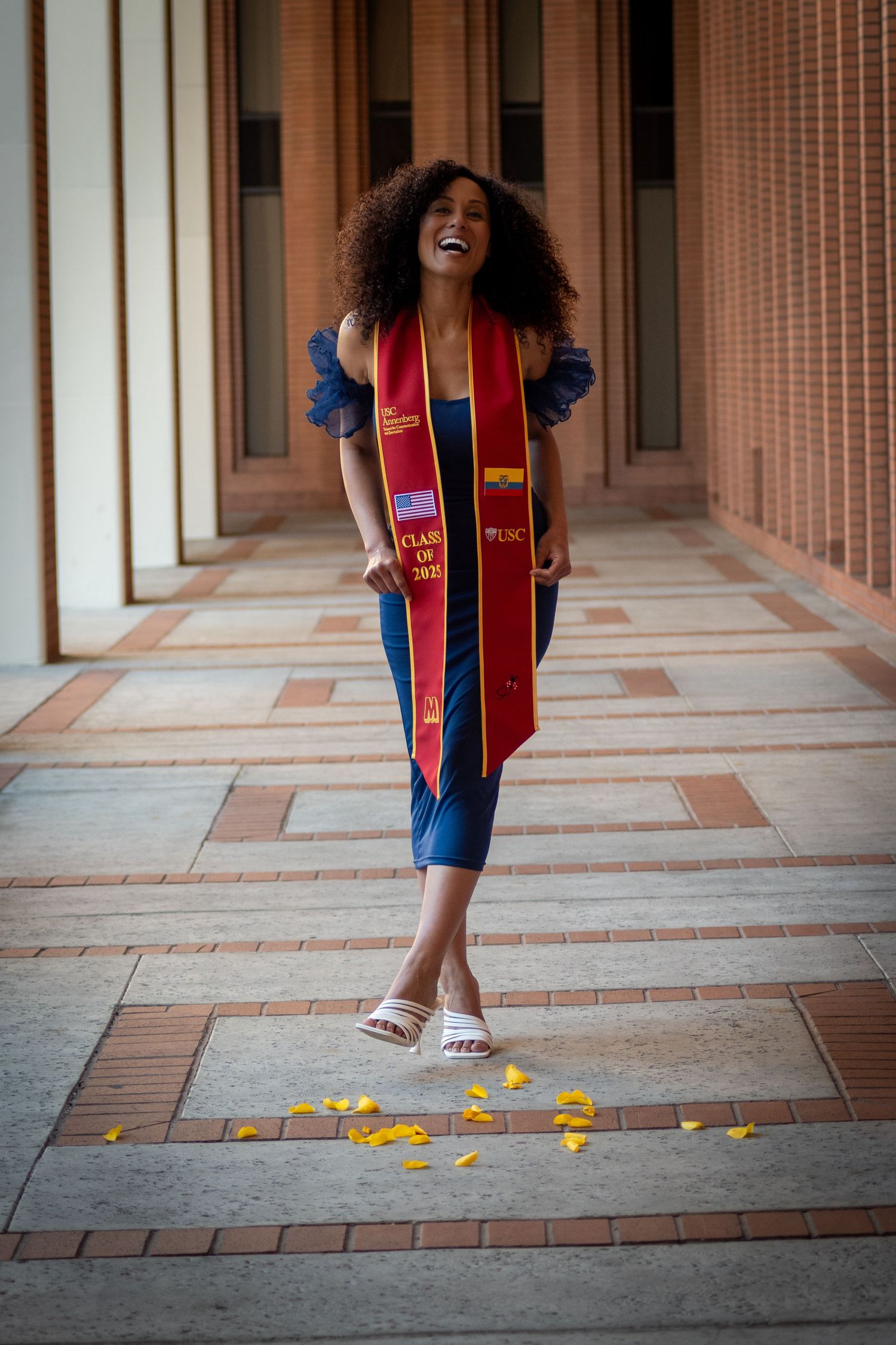 Graduate in blue dress and red graduation stole walks joyfully through campus corridor with scattered yellow petals.