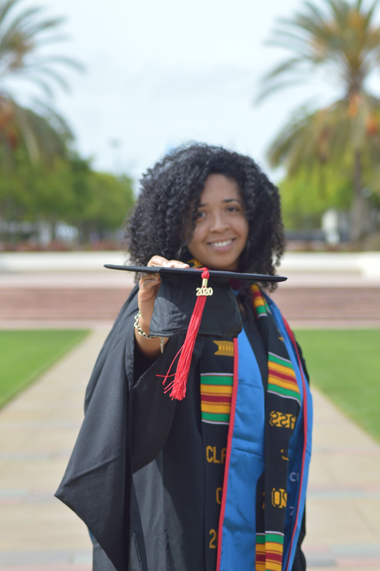 Graduate in black cap and gown with colorful kente stole celebrates outdoors with palm trees in background.