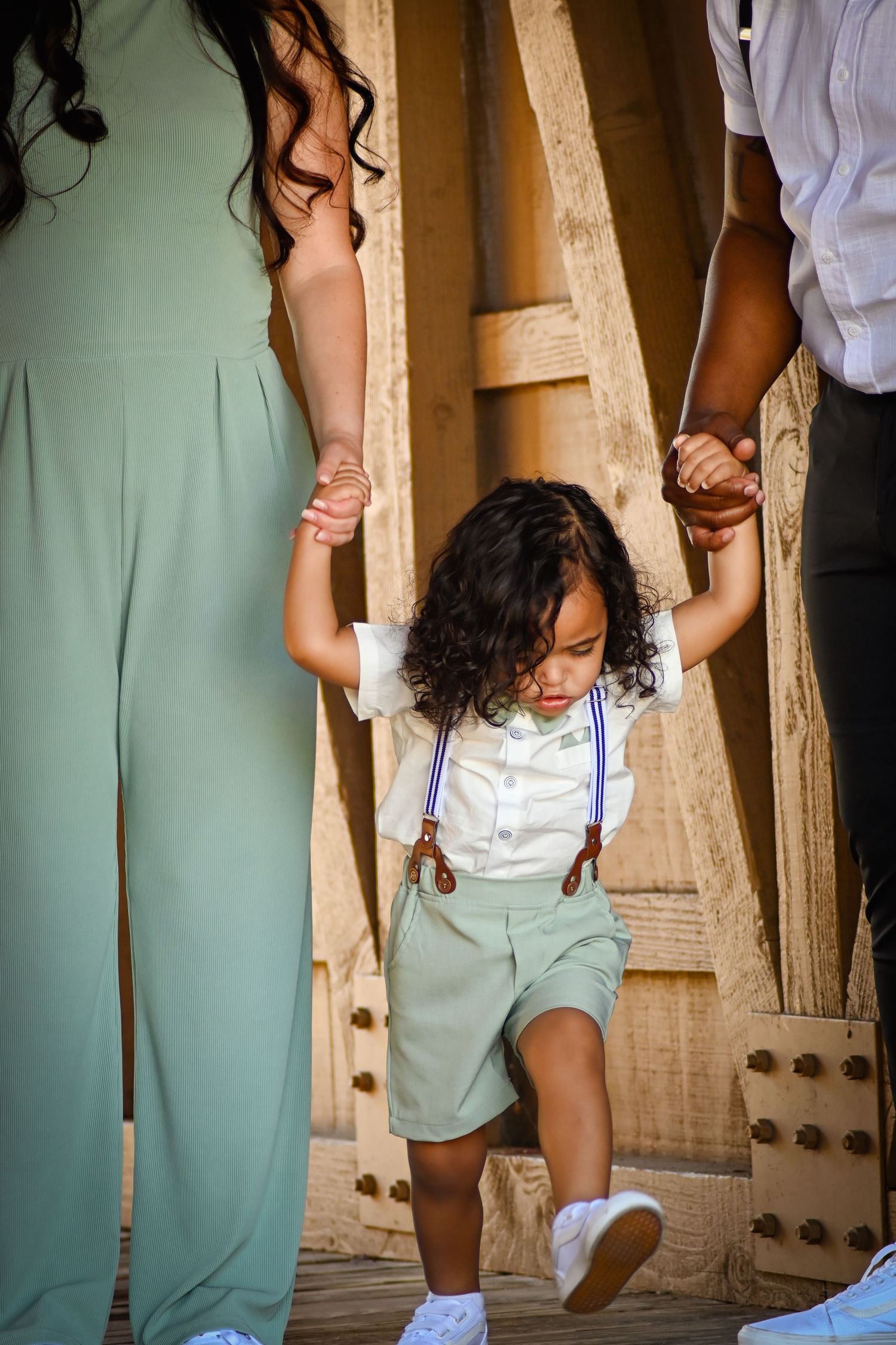 A child in mint green shorts and suspenders playfully swings while holding hands with adults in a rustic setting.