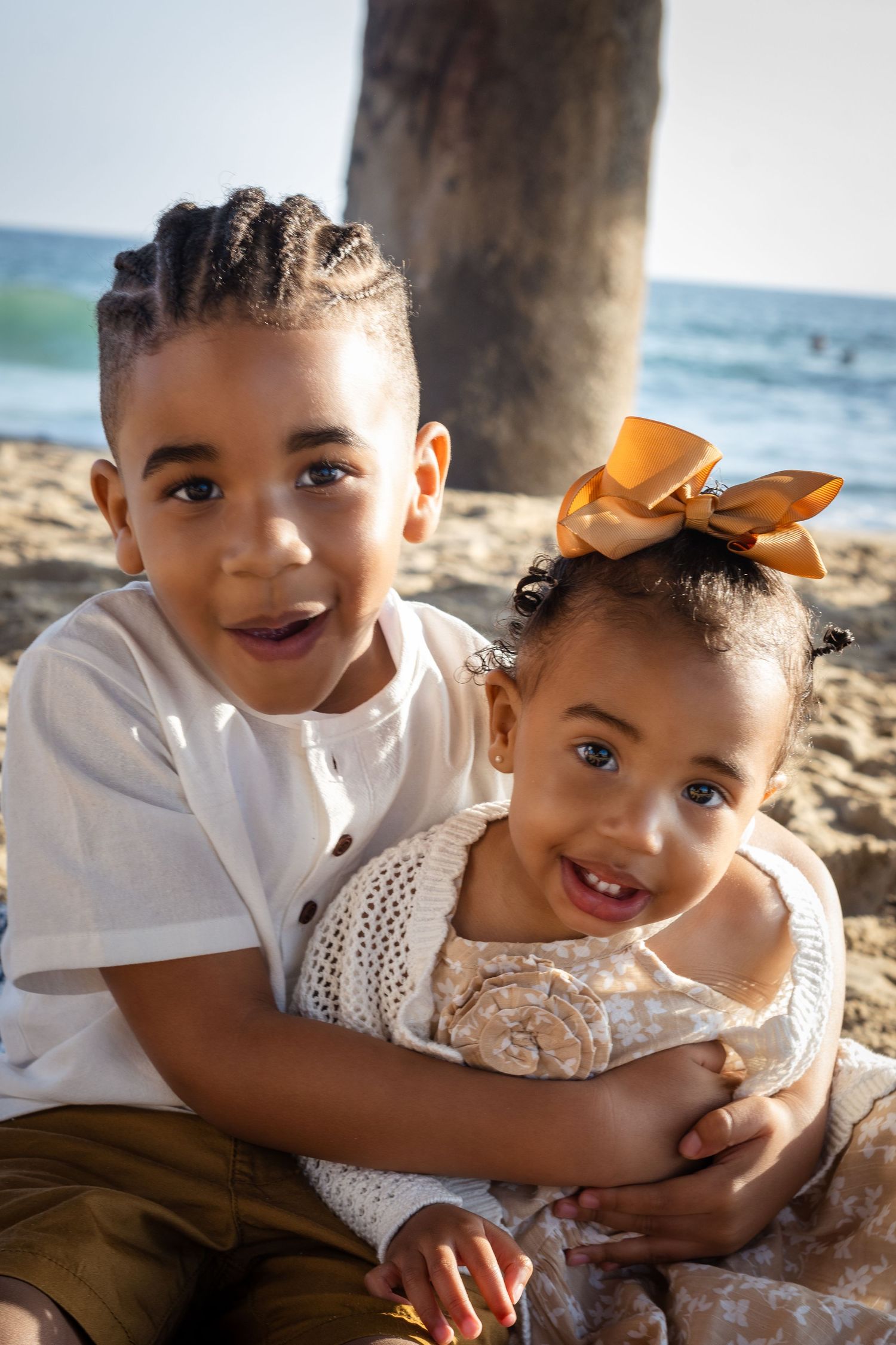 Two children sitting together on a beach smiling, with a wooden pier post in the background.