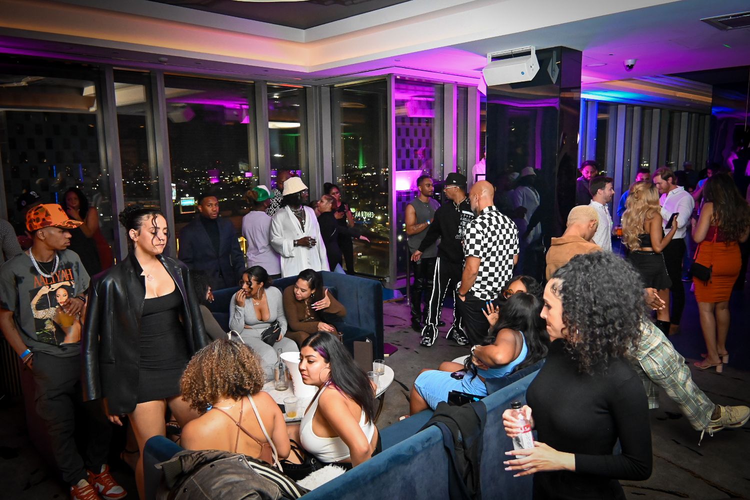 Nightclub crowd enjoying a party with purple and blue lighting illuminating the dance floor and bar area.