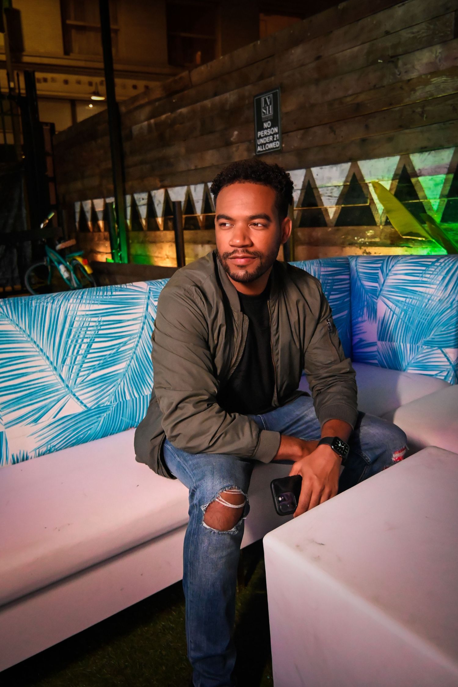 Person sits on white bench against brick wall with tropical neon palm leaf decor in background.
