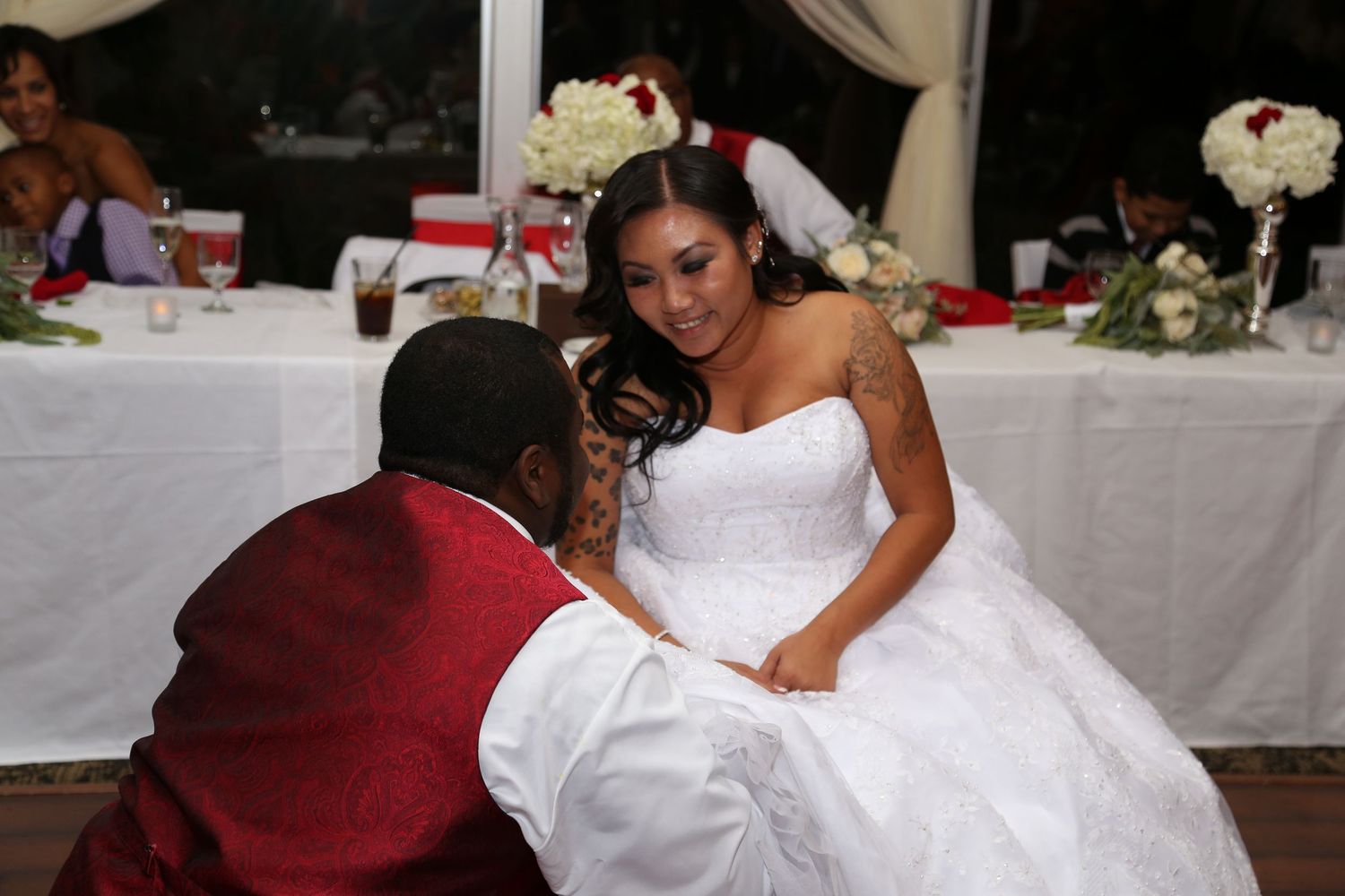 Groom kneels before bride in white ball gown during wedding reception ceremony.