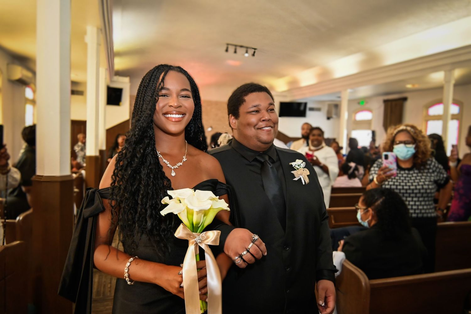 Smiling couple walks down church aisle, woman in black dress holding white calla lilies bouquet during wedding ceremony.