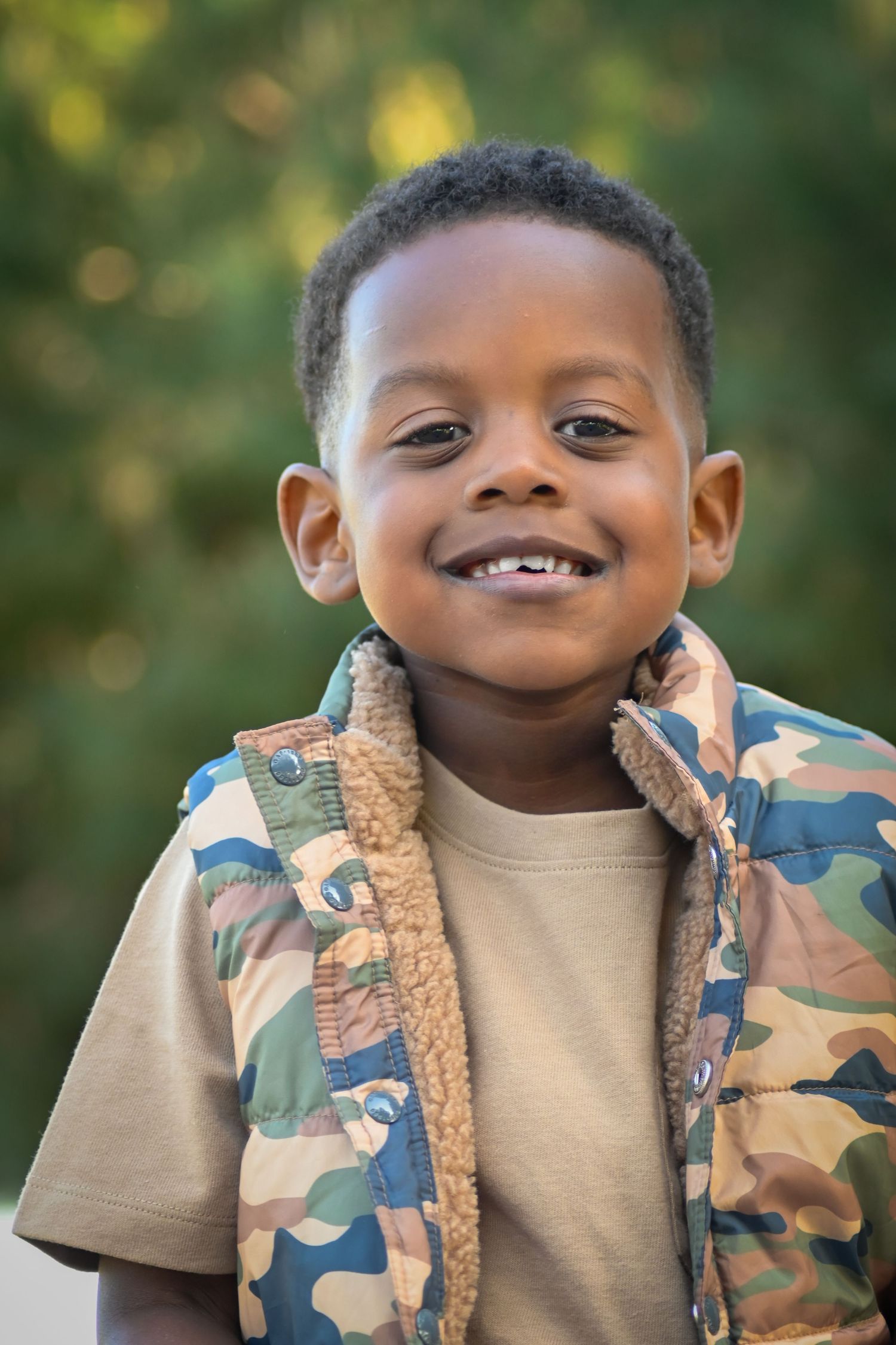 Smiling young Black boy wearing a camouflage vest outdoors with green trees in the background.