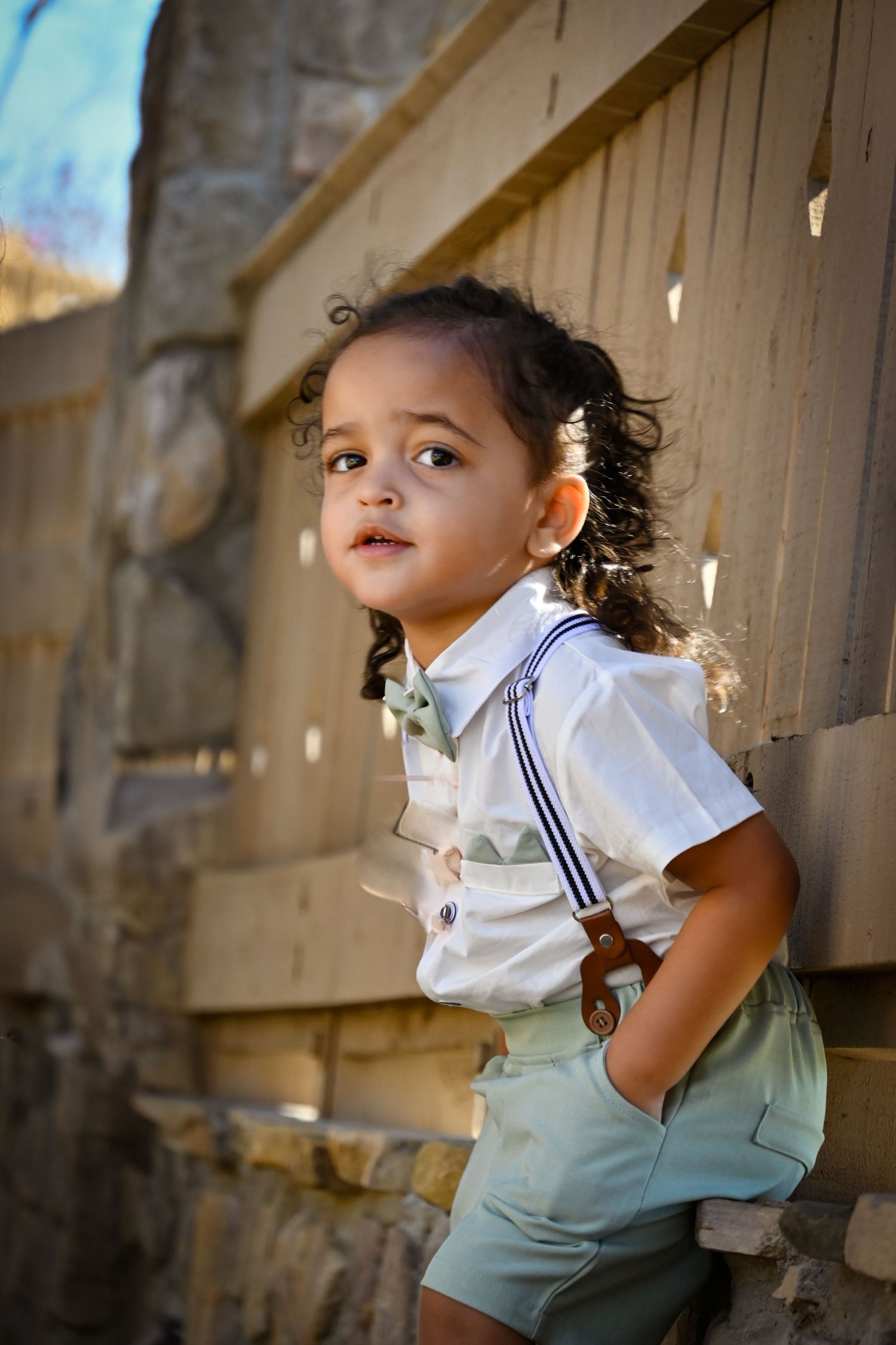 Adorable toddler in white shirt, suspenders, and green shorts leaning against a wooden fence outdoors.