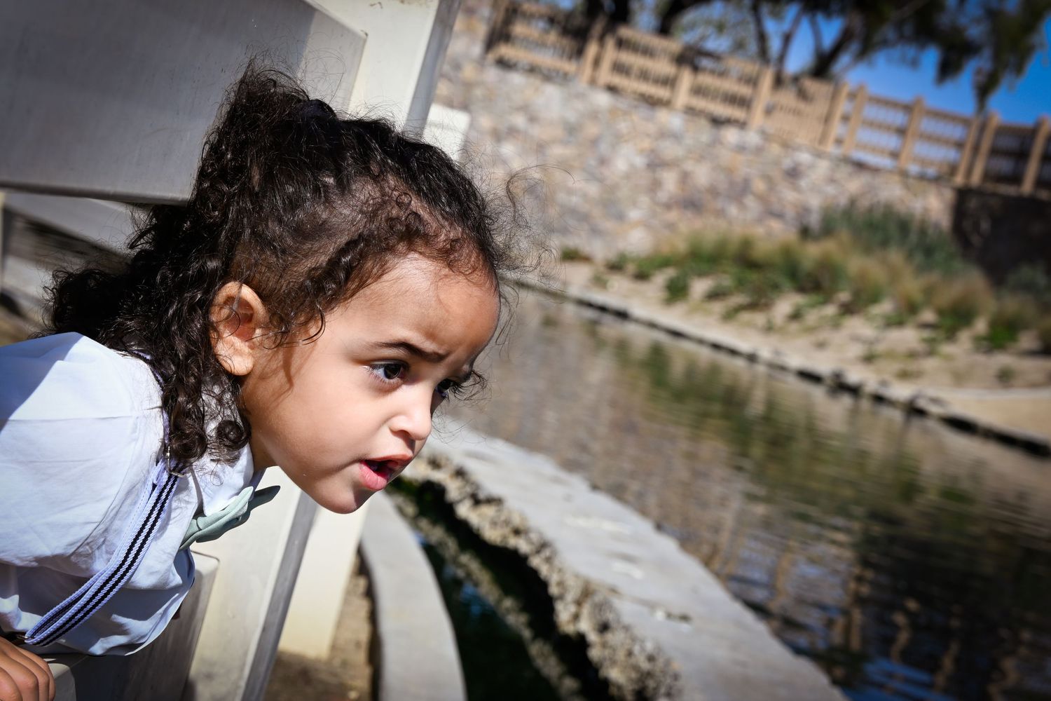 Young girl leaning over a railing, sticking out her tongue near a pond in an outdoor park setting.