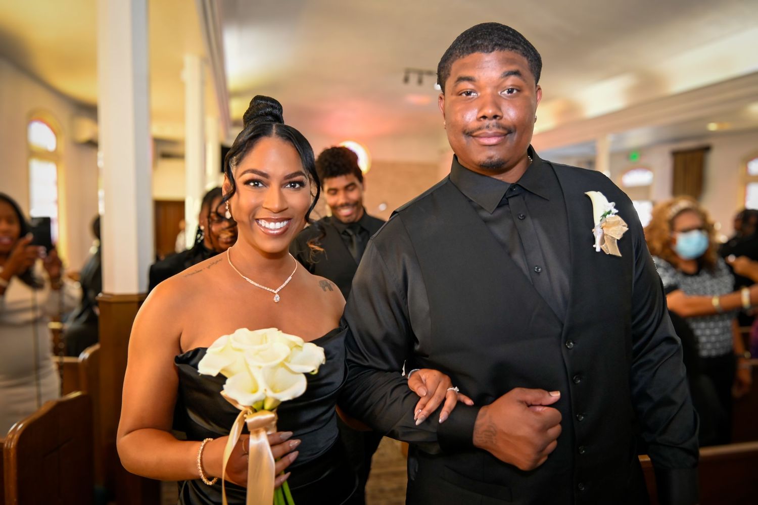 Smiling couple in black formal attire walking down church aisle, woman holding white roses bouquet.